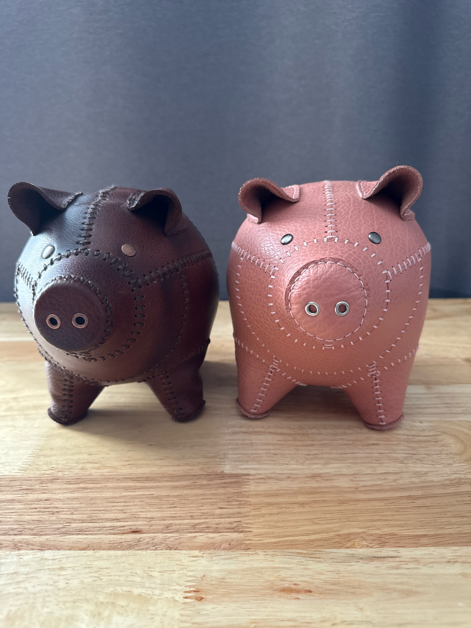 Two leather piggy banks, one brown and one pink, are positioned on a wooden surface against a dark gray background. The brown piggy bank is on the left, and the pink piggy bank is on the right. Both piggy banks are facing forward and are detailed with visible stitching and metal accents for eyes and nostrils. The wooden surface displays a natural wood grain pattern. The image does not provide any information about a location in New York City, United States of America.