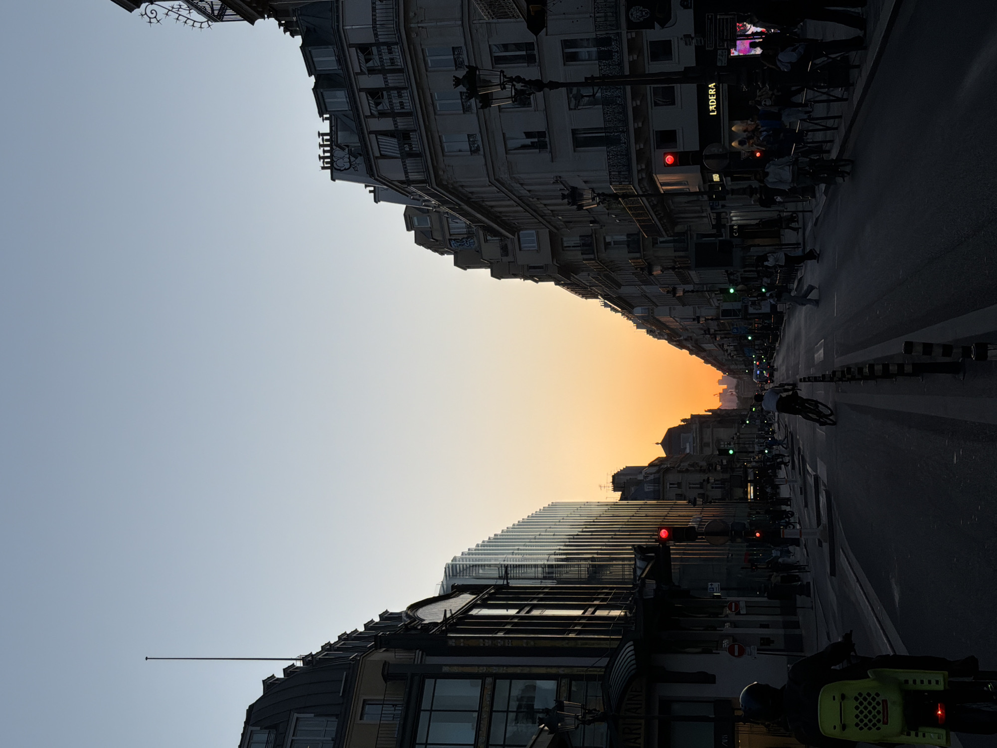 This image, captured at either dawn or dusk, depicts a bustling city street framed by tall buildings, with a warm, glowing sky between them. The street extends into the distance, creating a sense of depth and perspective.
On the left side of the street, a row of classic multi-story buildings features ornate balconies and detailed facades, indicative of traditional European architecture. On the opposing right side, a more modern building with numerous reflective windows creates a striking contrast.
The asphalt street is populated by several individuals. People are seen walking on the sidewalks and riding bicycles. One bicycle in the foreground is equipped with a distinctive bright yellow child seat or carrier. Multiple traffic lights are visible along the thoroughfare, displaying both red and green signals, suggesting active traffic control. Traditional street lamps are also present, illuminating parts of the scene.
The sky transitions from a soft blue-grey at the top to a vibrant orange-yellow nearer the horizon, characteristic of sunset or sunrise, casting long shadows and highlighting the silhouettes of the buildings and people. A sign with the word "LADERA" is visible on one of the buildings on the left side of the street. The overall scene conveys the serene yet active atmosphere of an urban environment during the golden hour.