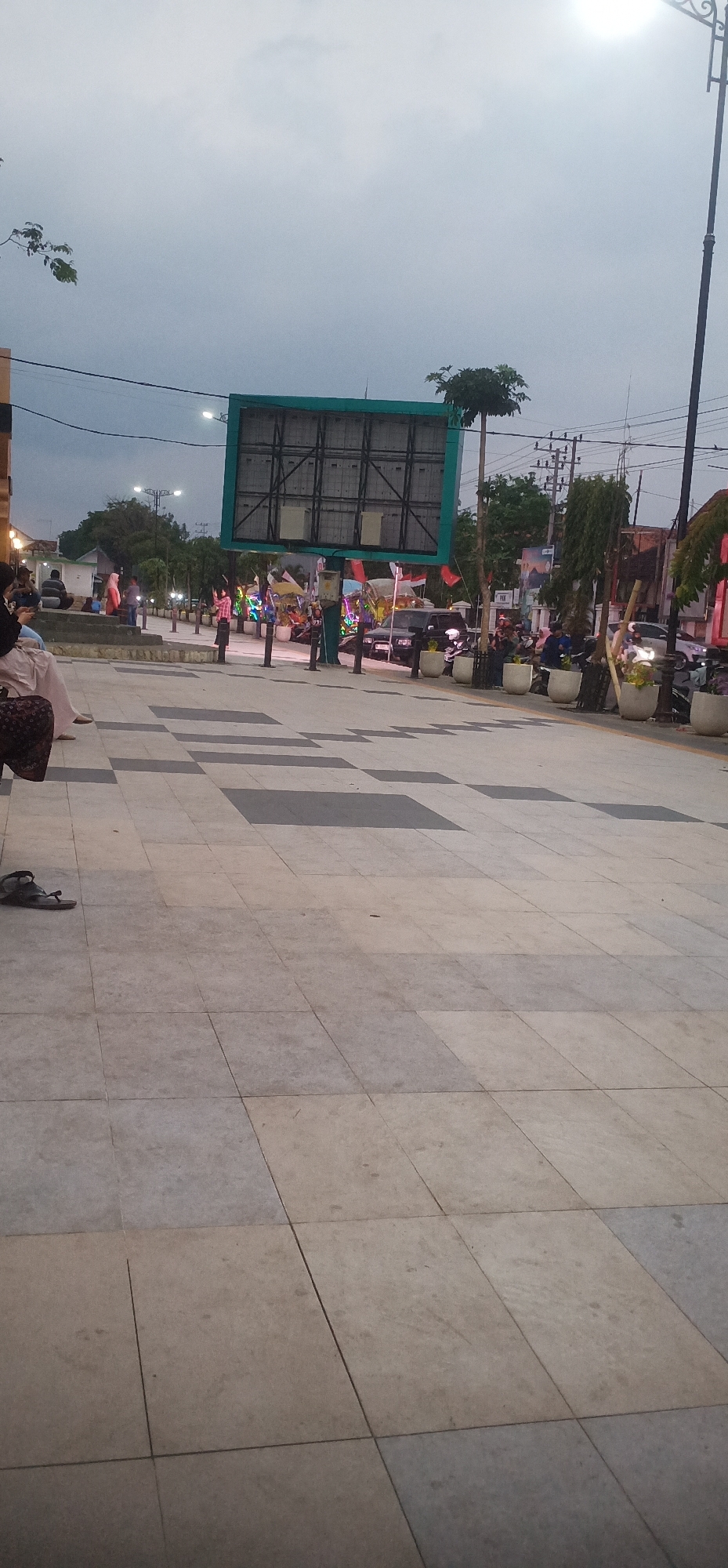 The image captures a street scene in Probolinggo, Indonesia, likely during dusk or a cloudy day. In the foreground, a tiled pedestrian walkway with a checkerboard pattern of light and dark gray tiles extends into the distance. A single sandal rests on the walkway near the bottom left.
In the mid-ground, a large, prominent billboard with a green frame and a dark, grid-like interior stands out. The billboard is positioned over a street or plaza area. To the left of the billboard, several people are seated on a raised concrete area, with some standing nearby. Further to the right, a street is visible with vehicles, including a dark SUV, and parked motorcycles. Along the street, decorative lights are illuminated, and several flags, including Indonesian flags, are displayed. Potted plants line the edge of the walkway.
In the background, trees and utility poles with overhead wires are visible against a cloudy sky. The overall impression is of a public space in a city, possibly a town square or a busy street, with people gathered and activities underway, suggested by the flags and lights.