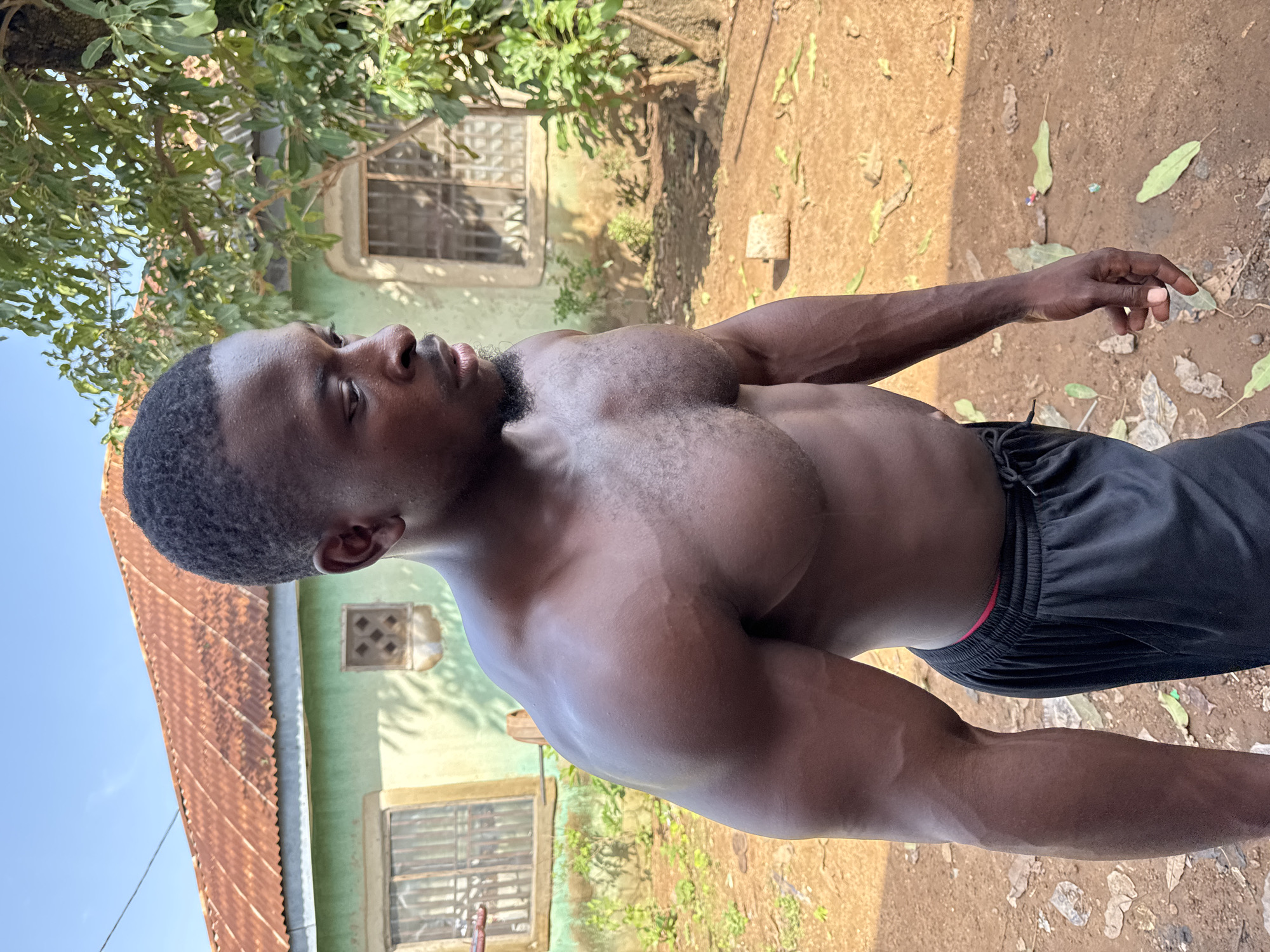 A muscular Black man stands outdoors, shirtless and wearing black shorts. He is positioned in the foreground, facing to the right with his head turned slightly. The background shows a light green building with a corrugated metal roof, a tree with green leaves, and a dirt ground covered with scattered leaves. The overall lighting suggests a sunny day. The location is Akwanga, Nigeria.