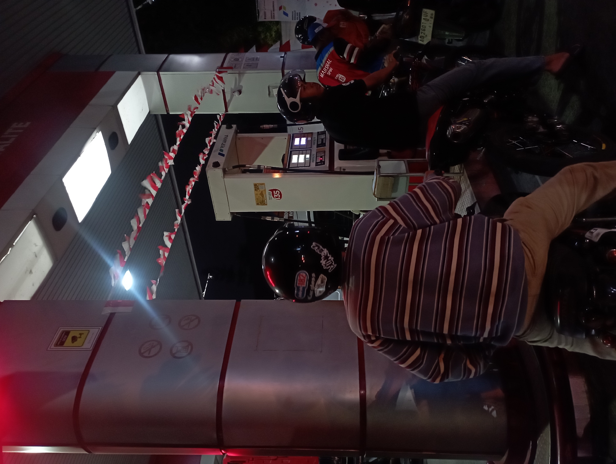 In Surakarta, Indonesia, at a Pertamina gas station at night, a group of people on motorcycles are present. The foreground features a person from behind, wearing a black helmet and a horizontally striped long-sleeved shirt in purple, white, and dark red, along with light brown pants. This individual is seated on a motorcycle. To the right, another person on a motorcycle is visible, wearing a black shirt and blue jeans, also wearing a helmet. Further right, a third person is seen wearing a red and blue vest over an orange shirt, also on a motorcycle. The gas station has multiple fuel pumps, one of which is prominently displayed with "PERTAMINA" signage. Red and white Indonesian flags are strung across the gas station canopy. Overhead lighting illuminates the scene, creating a nighttime ambiance. A license plate visible on a motorcycle reads "AD 2601 AUF".