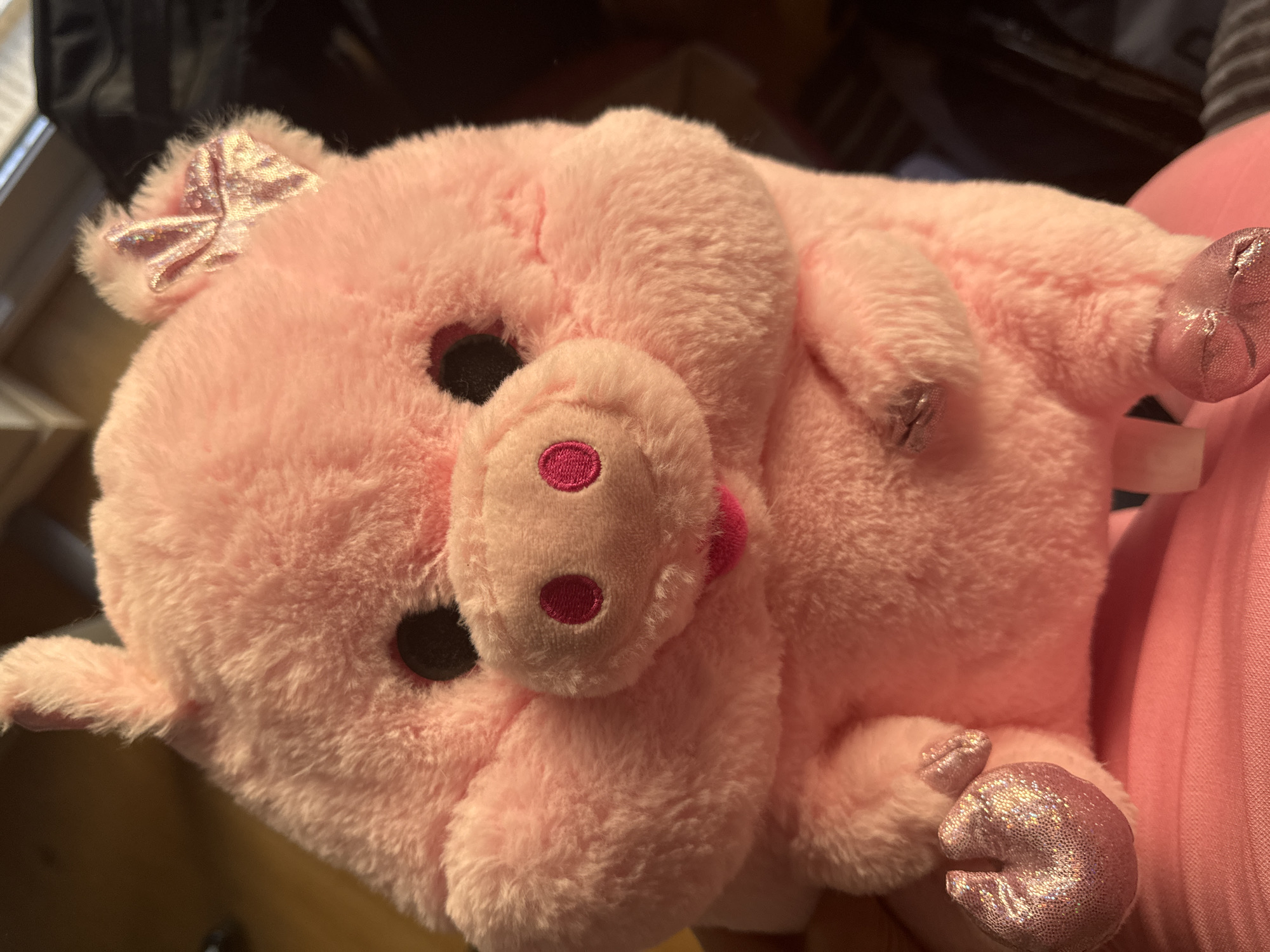 A close-up, high-angle shot focuses on a pink, plush pig toy. The toy has large, round, black eyes and a snout with two pink circles for nostrils. A small pink bow with glitter is attached to the pig's head, near its left ear. The pig's mouth is slightly open, revealing a pink tongue. Its cheeks are puffed out, giving it a chubby appearance. The pig's front feet are visible, each with two toes, and are adorned with glittery pink material. The toy is resting on a pink surface, possibly clothing or a blanket. The background is dark and blurred, with hints of wooden furniture and what appears to be a window with blinds. The lighting suggests an indoor setting, possibly a home. There is no indication of any specific location within the city of Murcia, Spain, in the image.
