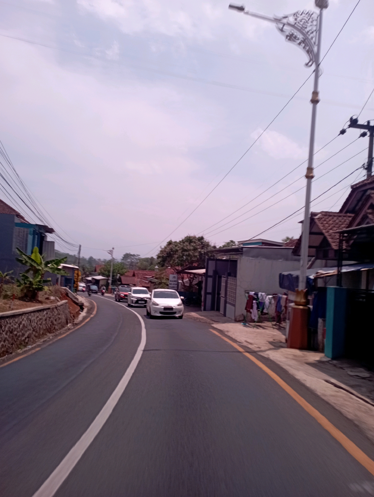 The image depicts a street scene in Payungsari, Indonesia, on a cloudy day. The road is paved and curves to the left, with a white line down the center and a yellow line along the right edge. Several cars are driving on the road, including a white sedan in the foreground on the right and another white sedan further down the road. On the left side of the road, there is a stone retaining wall topped with vegetation, and a building with a blue facade and a banana plant. On the right side of the road, there are houses with tiled roofs, power lines, and a street lamp with a decorative design. Clothes are hanging on a line in front of one of the houses. The sky is overcast with pale clouds.
