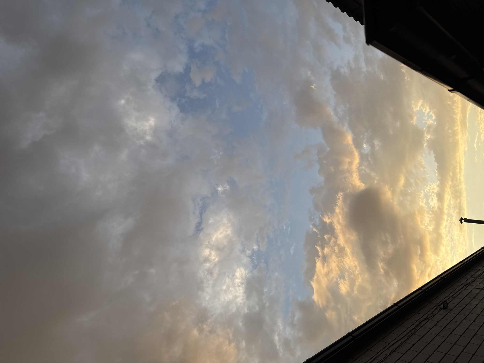 The image shows a sky with clouds at sunset, with the tops of two buildings visible in the foreground, one on the left and one on the right. The building on the left is a dark-colored house with a textured surface, possibly brick. The building on the right is darker and appears to be a roofline. A thin, dark chimney is visible extending from the house on the left. The sky is filled with a mix of gray and white clouds, with patches of orange and yellow light indicating the setting sun. Some areas of blue sky are visible between the clouds. The overall impression is of a cloudy sunset over a residential area. The location is specified as Mikhaylovsk, Russia.