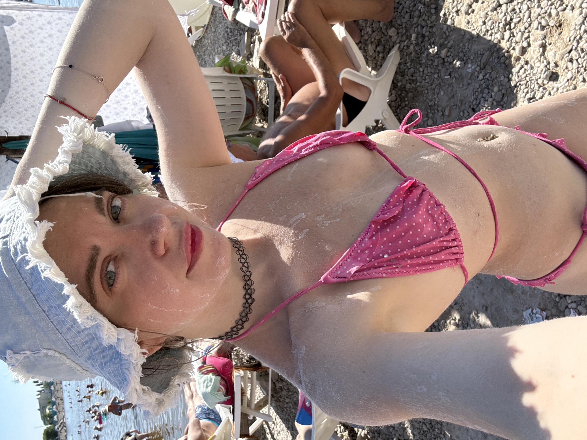 This media file features a young woman lying on a pebble beach in Sol-Iletsk, Russia. She is wearing a light blue denim bucket hat with frayed edges and a pink polka dot bikini. Her skin is dusted with what appears to be sand or sunscreen. The woman is looking upwards and slightly towards the camera, with a neutral expression.
In the background, other people are visible. One person is seated on a white plastic chair, with their legs extended. Another individual, partially obscured, appears to be wearing dark bottoms. Further back, a body of water, likely a lake or river, is visible with numerous people swimming or wading in it. Some are standing in the shallow water, while others are further out.
The scene suggests a sunny day at a beach or resort. The sunlight creates shadows on the ground, indicating it is daytime, likely midday or afternoon. The presence of people in swimwear and the beach setting point to a recreational activity, such as sunbathing or swimming. The overall mood is relaxed and summery. No visible text is present in the image.