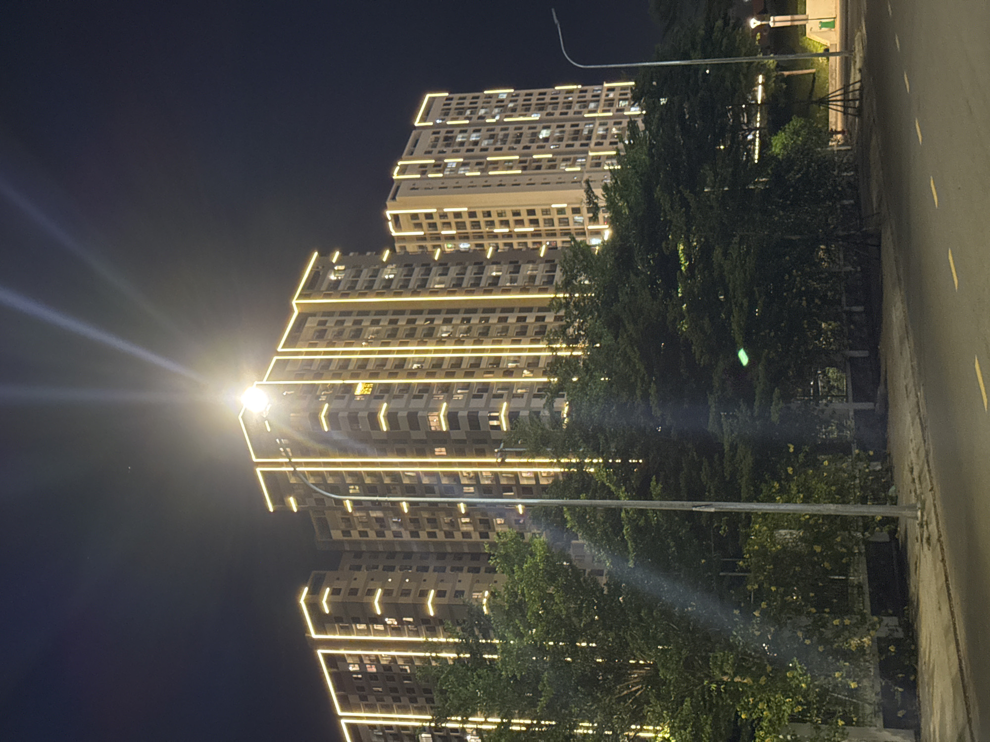A nighttime exterior shot captures a modern apartment complex in Đà Nẵng, Vietnam, illuminated by bright yellow exterior lighting. The tall, multi-story buildings are outlined with these lights, accentuating their architectural features. In the foreground, lush green trees and foliage obscure the lower portions of the buildings. A street lamp stands prominently in the center of the frame, casting its own light. A paved road runs across the bottom of the image, with yellow lane markings visible. The sky is a dark, inky black, typical of nighttime. The overall scene depicts an urban environment at night, highlighting the illuminated architecture.