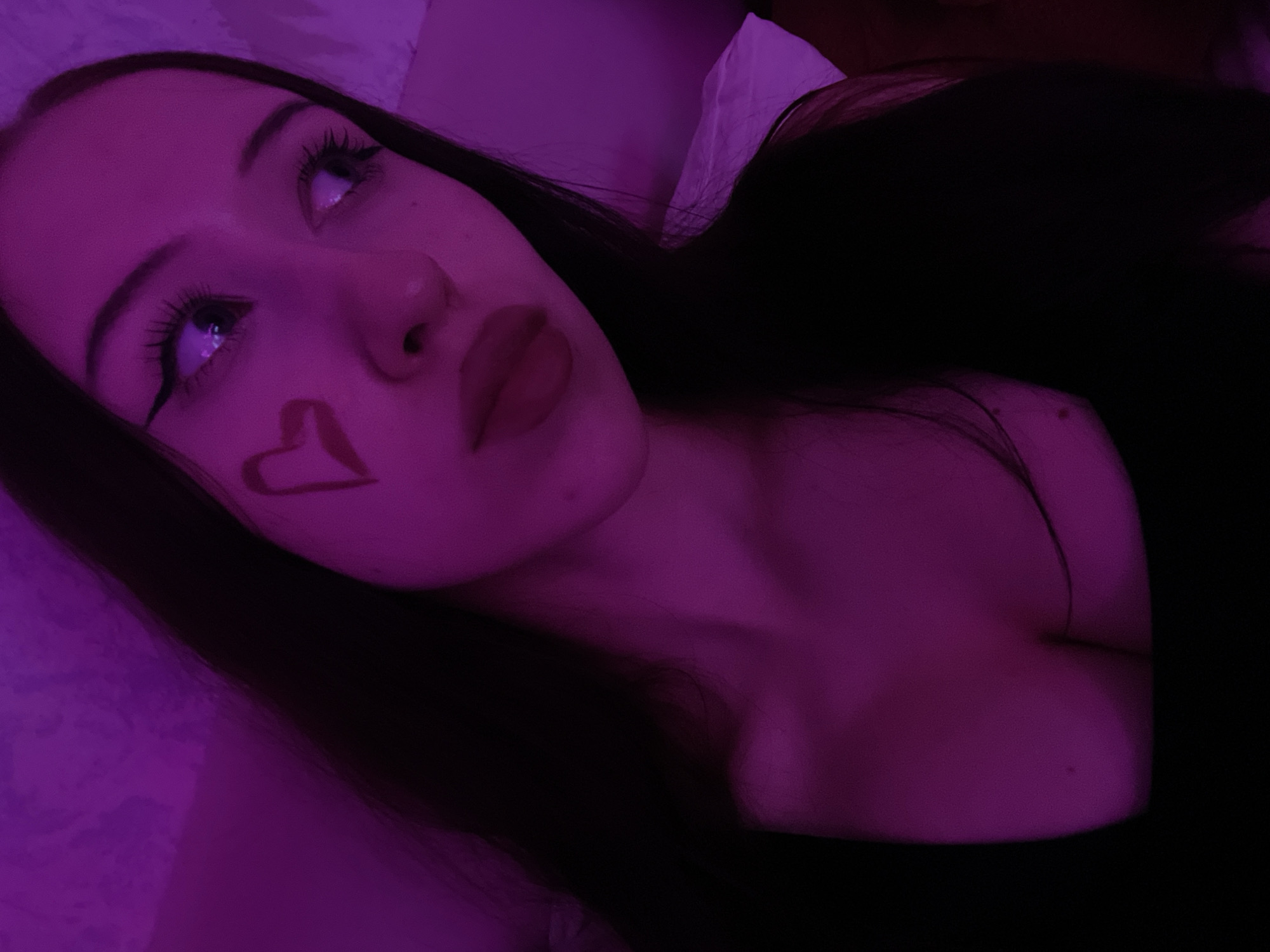 This is a close-up, medium shot portrait of a young woman with long, dark hair. She is looking to her right, away from the camera. She is wearing dark clothing and has a drawn-on heart with red lipstick on her left cheek. Her eyes are open and her eyelashes are prominent. The lighting in the image is predominantly purple. The background appears to be a wall or fabric that is also illuminated with purple light. The setting is indoors. There is no indication of the location in Votkinsk, Russia.