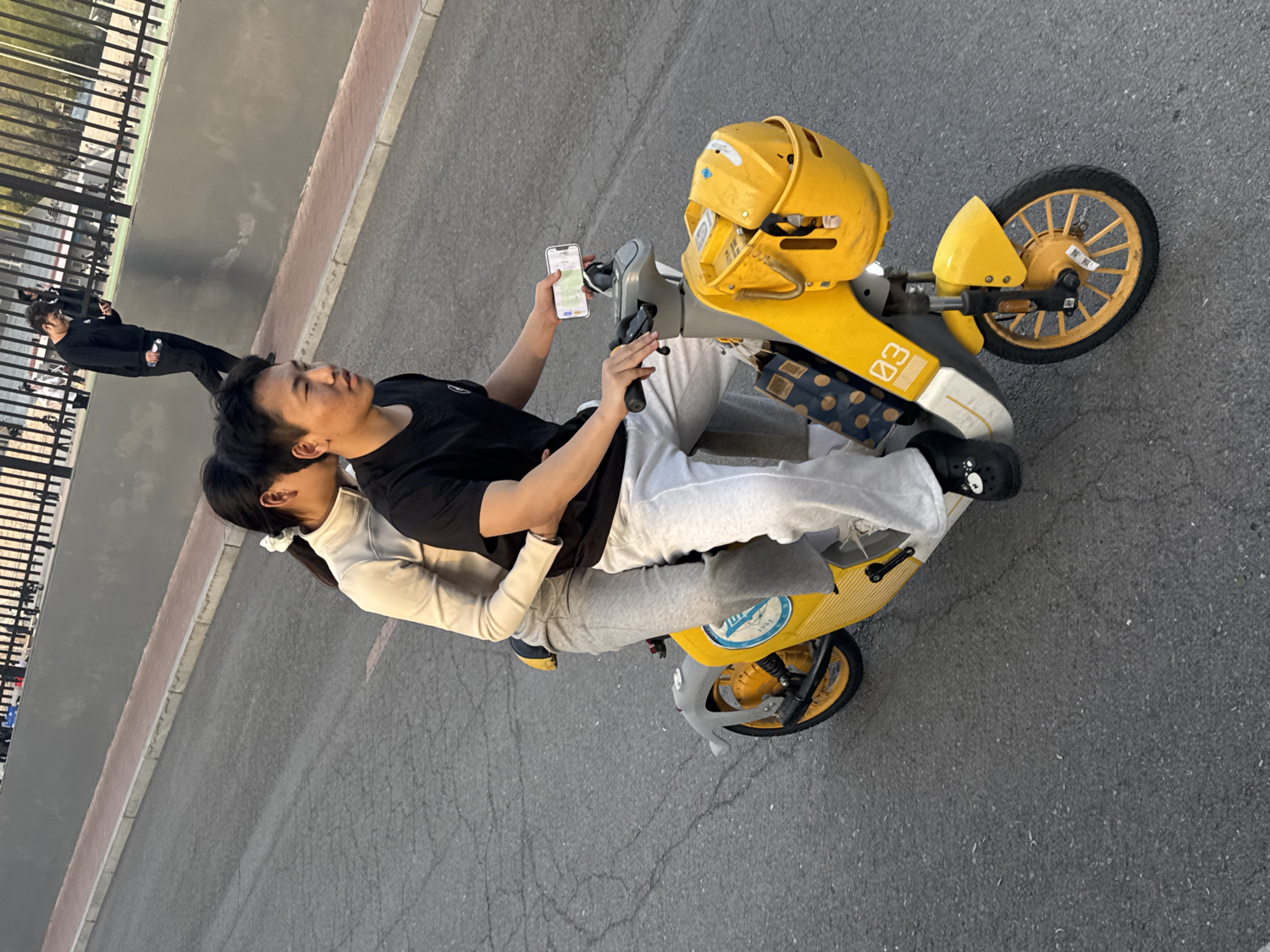 The image shows a man and a woman riding a bright yellow electric scooter on an asphalt road in Zhongtun, China. The man, seated in front, wears a black t-shirt and light gray sweatpants, with black slip-on shoes adorned with white bear faces. He holds a smartphone in his right hand, which displays a map or navigation app. The woman is seated directly behind him, wearing a light-colored long-sleeved top and light gray sweatpants, with her arms wrapped around his waist.
The scooter itself is predominantly yellow with grey accents. Two yellow helmets are visible, one resting on the handlebars, which has the number "03" written on it. The side of the scooter also features the number "03" in white text. A partially visible blue circular logo near the base of the scooter contains the number "1954".
In the background, another person dressed in dark clothing walks along a path bordered by a black metal fence and a low concrete wall. The setting appears to be an outdoor urban or suburban area during daylight hours. The activity suggests the couple is either navigating or preparing for a ride on the shared electric scooter.