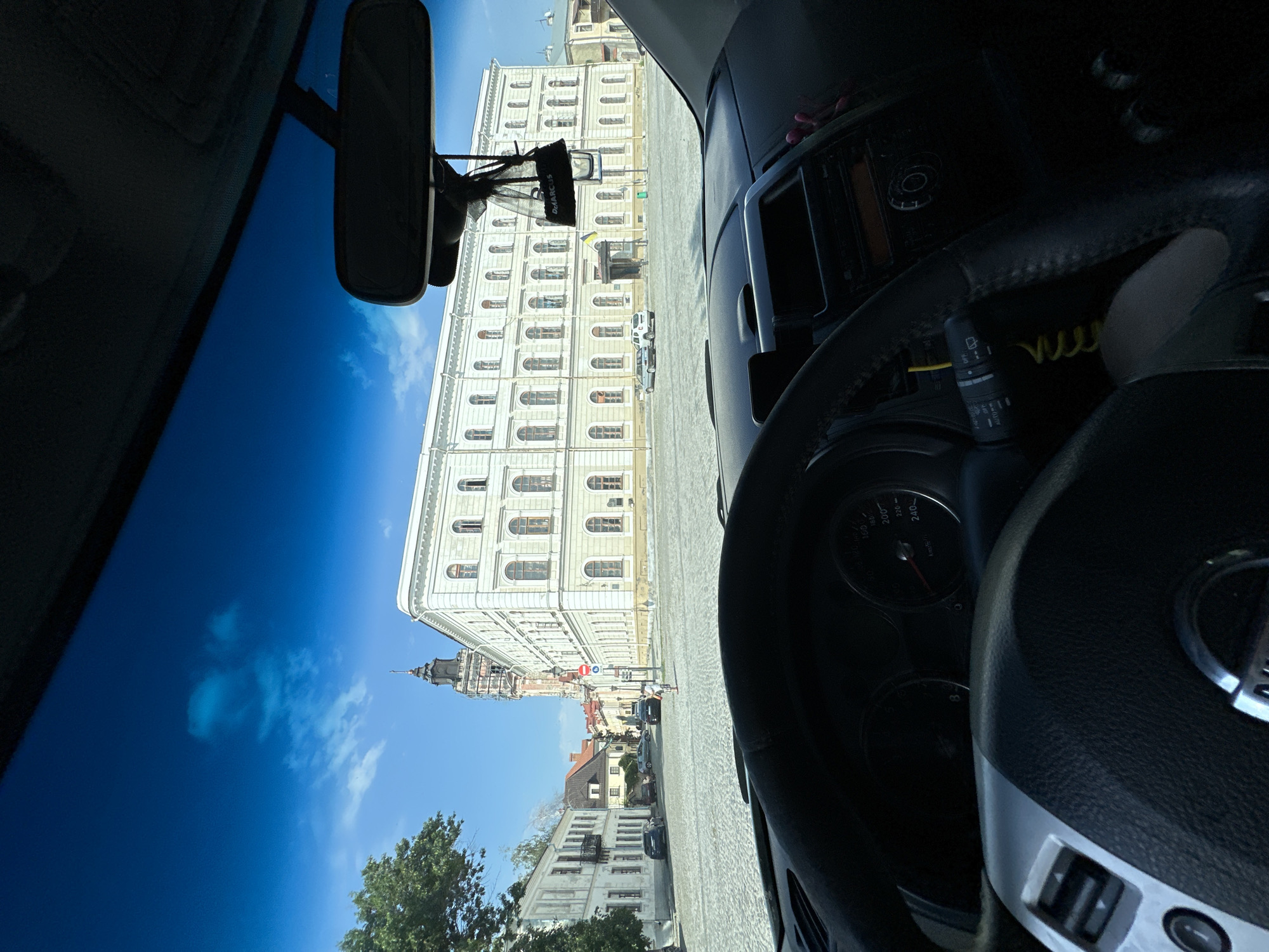 The image captures a view from inside a car, looking out through the windshield. The car is parked on a cobblestone street in Kamianets-Podilskyi, Ukraine, facing a large, pale building with many windows and a tall tower with a dark spire. The sky is bright blue with a few scattered clouds.
In the foreground, the car's dashboard and steering wheel are visible. The steering wheel is black leather with silver accents. To the right of the steering wheel, part of the car's console is visible, including air vents and possibly a radio. A rear-view mirror hangs from the windshield, with a small black car air freshener dangling from it.
The street appears to be a public square or plaza, paved with cobblestones. Several cars are parked on the street, and a person, appearing to be a child, is standing in the middle of the square. The architecture of the surrounding buildings suggests a historic European city. The large building directly ahead is imposing and has classical architectural elements. The tower is a prominent feature of the skyline. The overall impression is of a sunny day in a picturesque, historic urban setting.