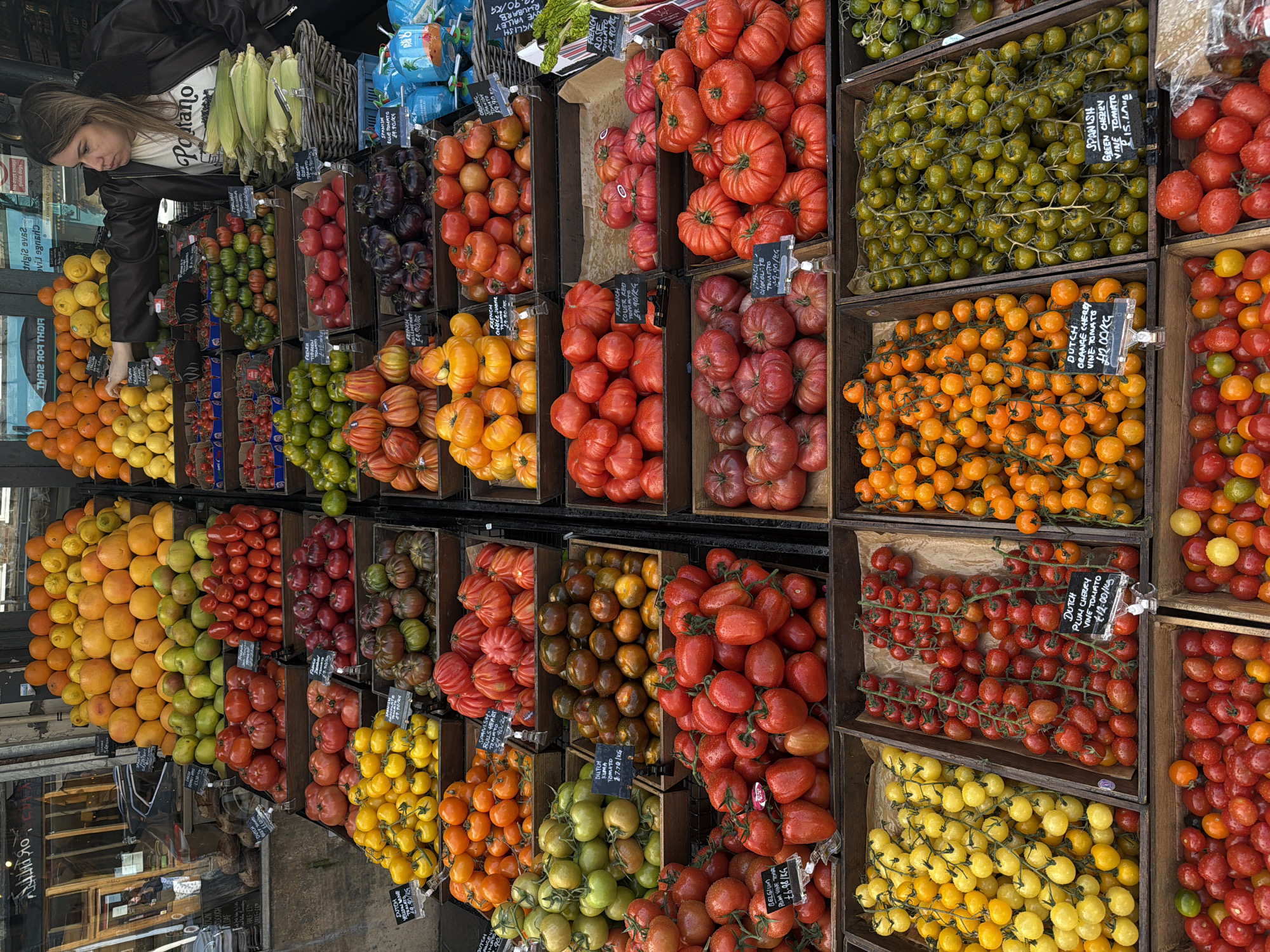 Here is a detailed description of the image:
**Scene:** The image depicts a vendor's stall selling a variety of tomatoes, citrus fruits, and corn on the cob. There are different tomato varieties, arranged in wooden crates, including orange cherry, green cherry, plum cherry, rose, bull's heart, chocolate, blue, yellow and common red tomato types. Each crate has a price tag. Oranges, lemons, and other citrus fruits are also displayed. The corn on the cob is stored at the back, along with containers that are unidentifiable.
**People:** A woman is visible in the image, looking at the fruit. She is wearing a black leather jacket and a t-shirt with a printed design.
**Location:** The location is London, United Kingdom, and based on the text visible in the image "LOAF", the location is likely Borough Market. The setting suggests an outdoor market environment.