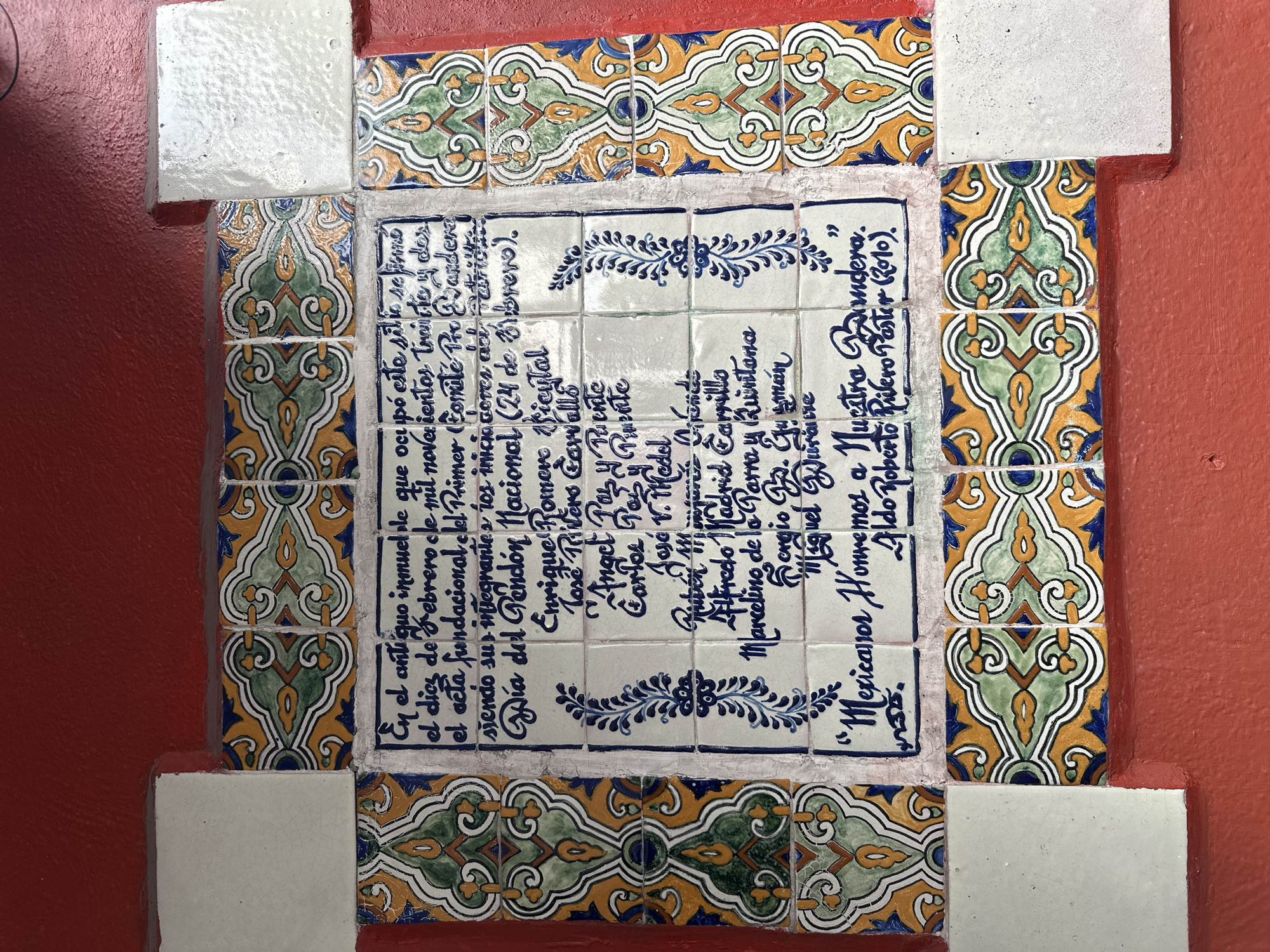 Here is a detailed description of the image:
The image shows a collection of decorative tiles set into a wall. The wall is painted a solid red color. The tiles in the center form a square containing text in blue ink, surrounded by blue decorative elements. The text is in Spanish, and appears to commemorate the founding of a committee on February 12, 1932, and includes a list of names. Surrounding this center square of tiled text is a decorative border of colorful tiles. These tiles have a repeating pattern in yellow, green, and blue. The entire tiled area is framed by sections of plain white tiles, which are set into the wall to create a border.
I am unable to provide a location in the city of Unknown, Unknown, as that information is not available in the text or image.