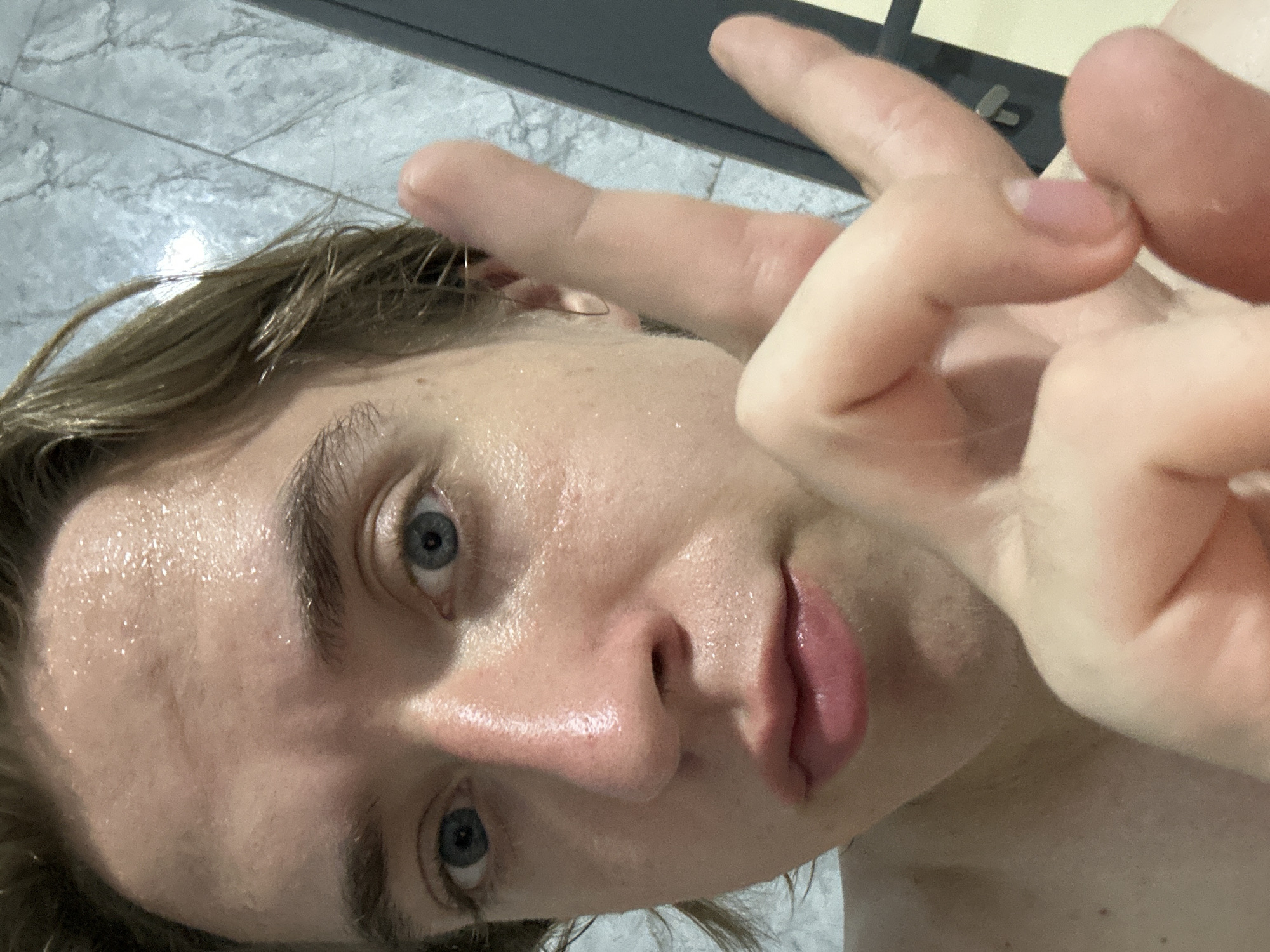 The image is a close-up, selfie-style photograph of a young Caucasian individual with light skin, fair hair, and blue eyes. The individual's forehead and face are visibly shiny with sweat, suggesting a warm or humid environment, or recent physical activity. Their hair is wet and damp, also indicating a similar condition. The individual is making a "peace sign" gesture with their right hand, with the fingers extended and the thumb tucked in. The background appears to be a tiled surface, possibly a bathroom or shower area, with a darker vertical element on the right side, which could be a door frame or cabinet. There is no discernible information about the location in An Hữu, Vietnam.