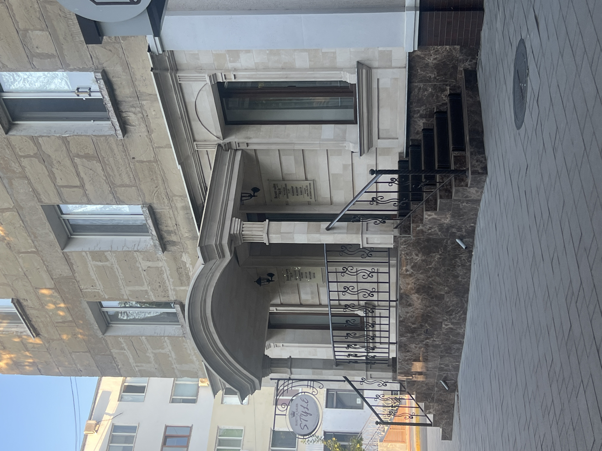The image shows the exterior of a beauty salon named "STYLE" located in Bender, Moldova. The building is constructed with light-colored stone blocks and features a prominent arched entrance with columns. A black wrought-iron fence lines the stairs leading up to the entrance. A sign for the salon is hanging to the left of the entrance, displaying the salon's name and a small crown emblem. A plaque with text is mounted on the wall near the entrance. The sidewalk in front of the building is paved with grey rectangular tiles, and a manhole cover is visible in the foreground. The building is situated next to a white residential building with multiple windows. The scene appears to be captured during daylight, with sunlight casting shadows on the building.