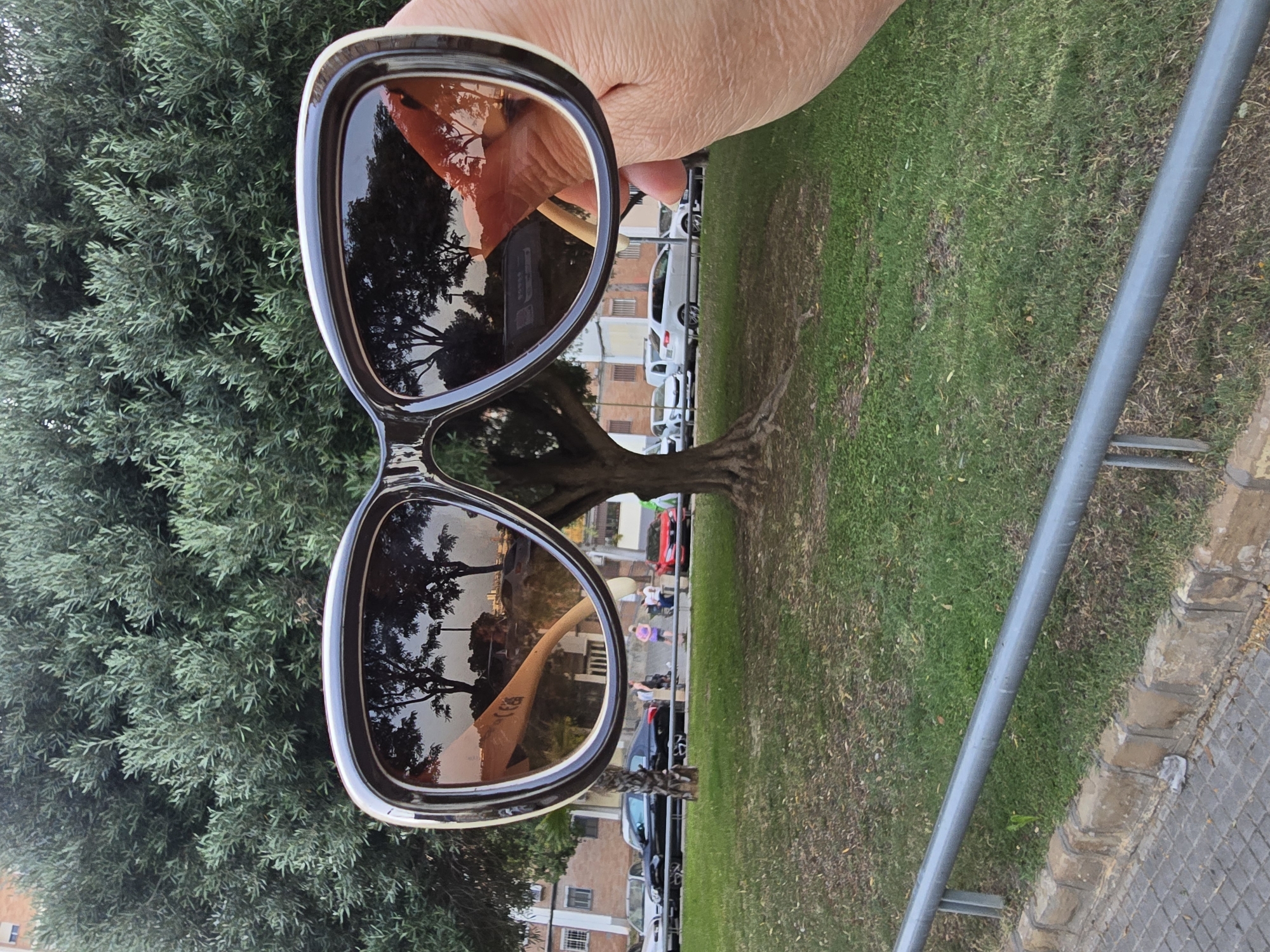 A pair of large, dark-lensed sunglasses are held up in the foreground, reflecting a tree-lined street with buildings and cars in the background. The reflection also shows a person's hand holding the sunglasses. The scene appears to be outdoors, possibly in a park or along a street, with grass and trees. The sunglasses have a white and dark brown frame. In the background, several cars are parked along the street, including a black sedan, a white SUV, and a red car. A few people are visible walking on the sidewalk. The overall lighting suggests it is either early morning or late afternoon. The location is identified as Cornellà de Llobregat, Spain.