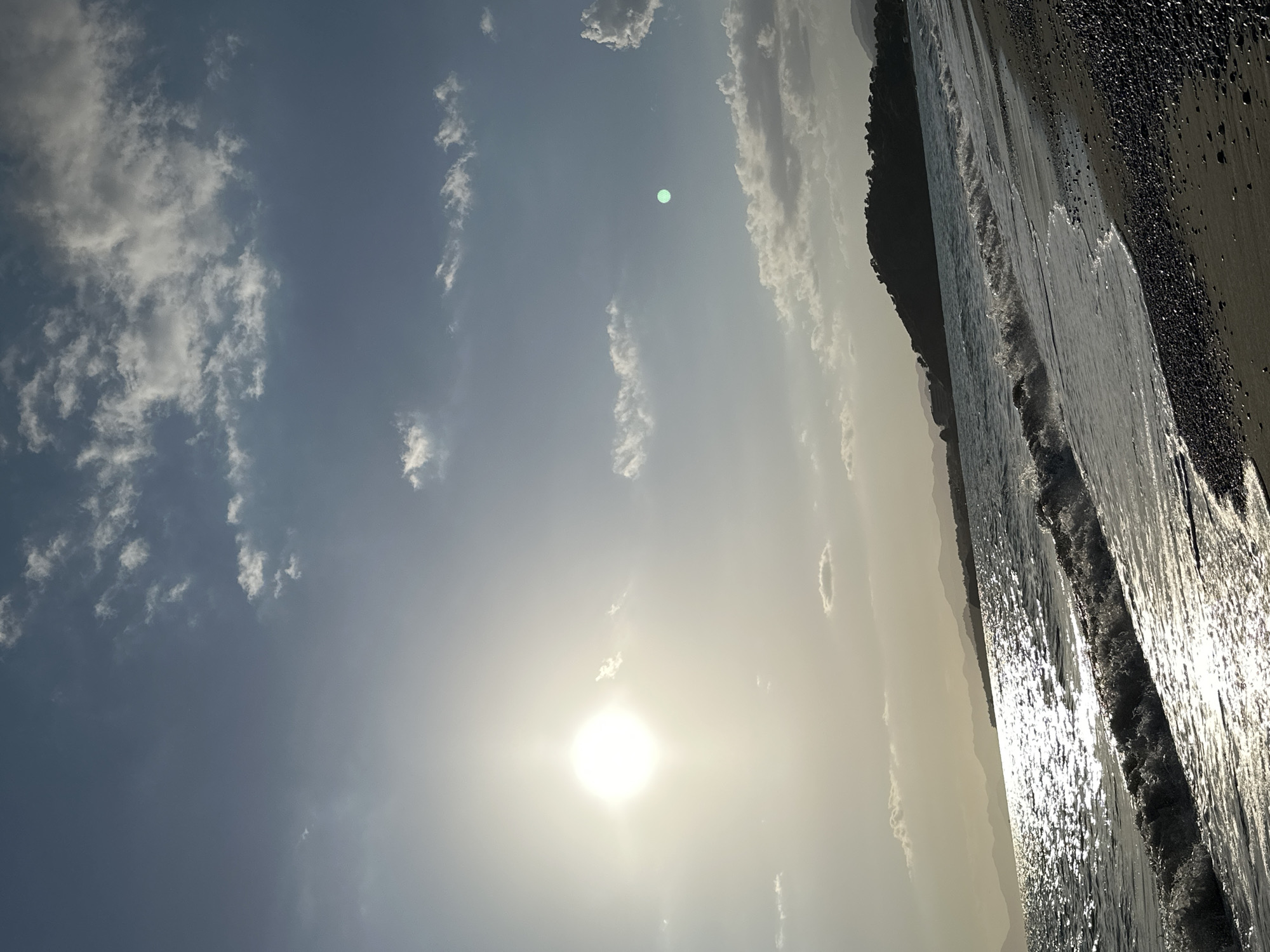 The image shows a beach scene in Fethiye, Turkey on a sunny day. The sky is light blue with scattered white clouds. The sun is bright and low in the sky, creating a glare. To the right of the sky, there is the coastline. The sea is calm with small waves lapping against the shore. The beach is dark, with what appear to be small rocks or pebbles. The waves are reflecting the sunlight. In the background, a dark landmass is visible. A green spot is present in the sky, possible lens flare.
