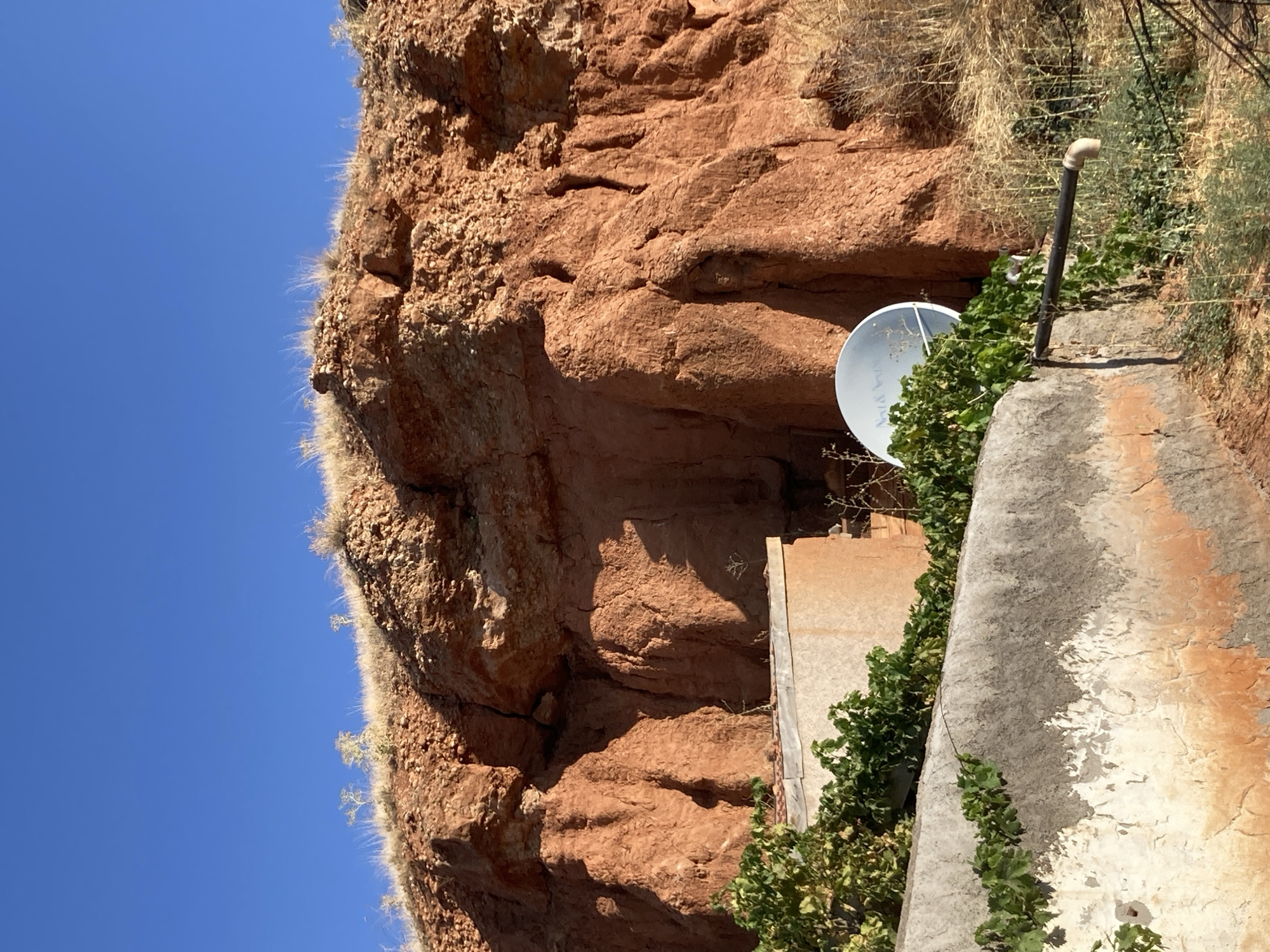 A satellite dish is attached to a concrete structure on a hill under a clear blue sky in Avanos İlçe Merkezi, Turkey. The satellite dish is white with a logo on it that reads "TV & Verve". The concrete structure is weathered and has green vines growing on it. Behind the concrete structure is a steep, red-rock cliff with sparse vegetation. The cliff has a rough, textured surface and appears to be eroded. The overall scene depicts a rural or semi-rural landscape with a mixture of man-made and natural elements.