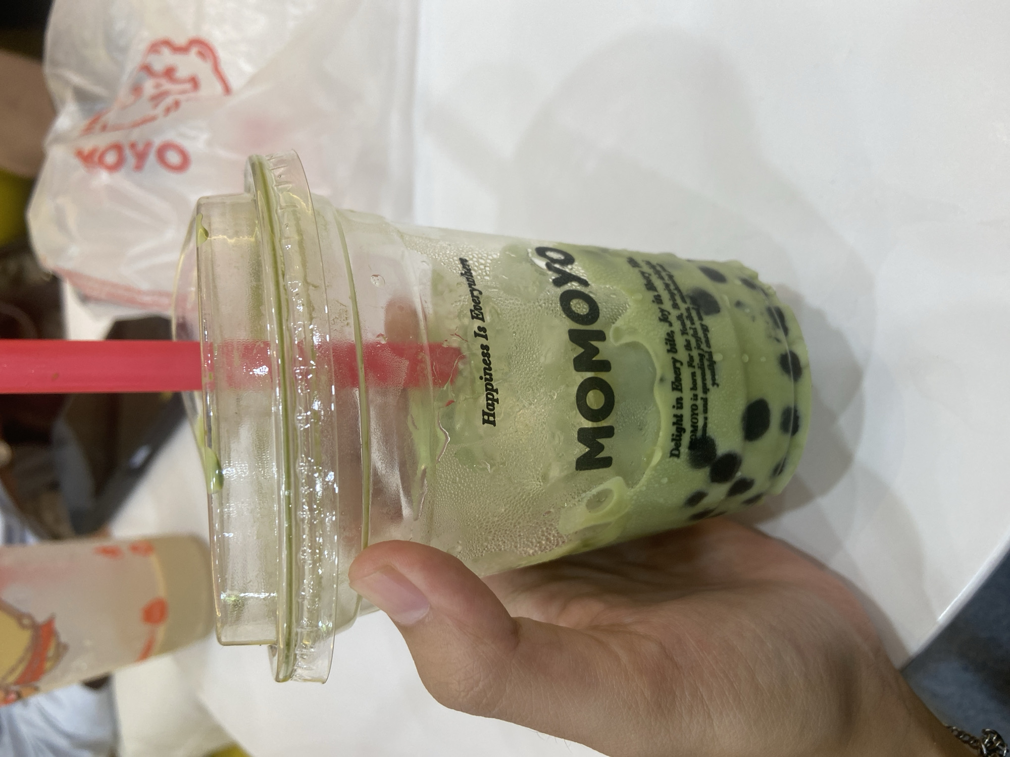 A close-up, slightly high-angle shot shows a hand holding a clear plastic cup filled with a green beverage and black tapioca pearls, commonly known as bubble tea. The cup has a clear plastic lid and a bright pink straw. The beverage has condensation on the outside of the cup, indicating it is cold. Text is printed on the cup, with the word "MOMOYO" prominently displayed in large, dark letters. Above "MOMOYO," smaller text reads "Happiness Is Everywhere," and below, additional text is partially visible, starting with "Delight in Every bite, Joy in Every." In the background, out of focus, is another clear plastic cup with a light-colored beverage and red accents, and a white plastic bag with red text and a graphic. The scene appears to be indoors, likely at a food establishment, with a white table surface in the foreground. There is no specific information about Kembang Harum, Indonesia present in the image.