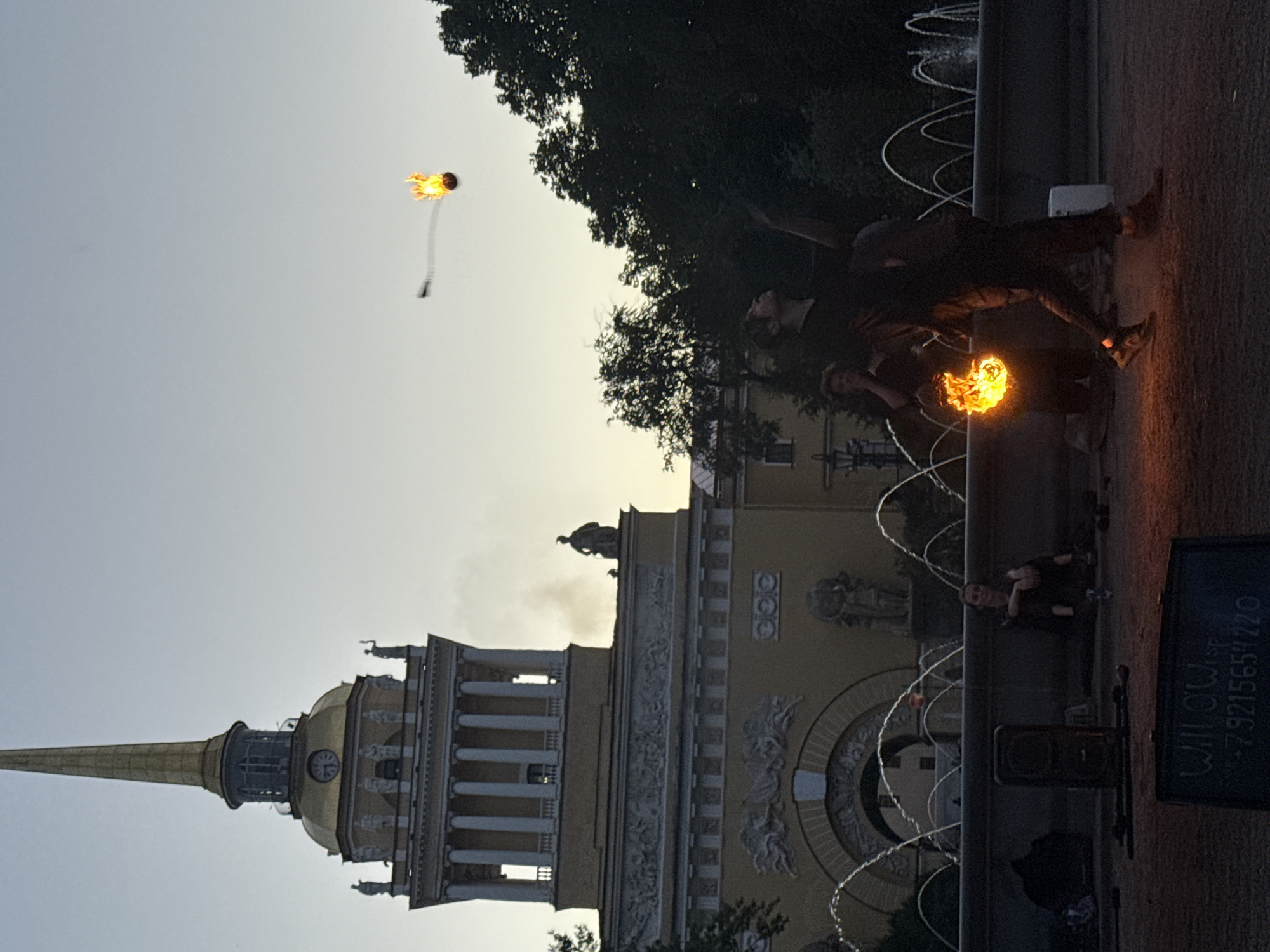 The image depicts a scene in Palace Square, Saint Petersburg, Russia, at dusk. A man is performing a fire-spinning act, juggling a flaming object in the air. He is wearing dark clothing and appears to be in motion. Behind him, a woman is seated by a fountain, and another person is visible in the background. The Admiralty building, with its distinctive golden spire, forms the backdrop. The sky is a pale blue, transitioning to a lighter hue near the horizon. The fountain is active, with arcs of water visible. A sign with the text "Willowisp" and a phone number is partially visible in the foreground. The overall atmosphere suggests an outdoor evening performance or gathering.