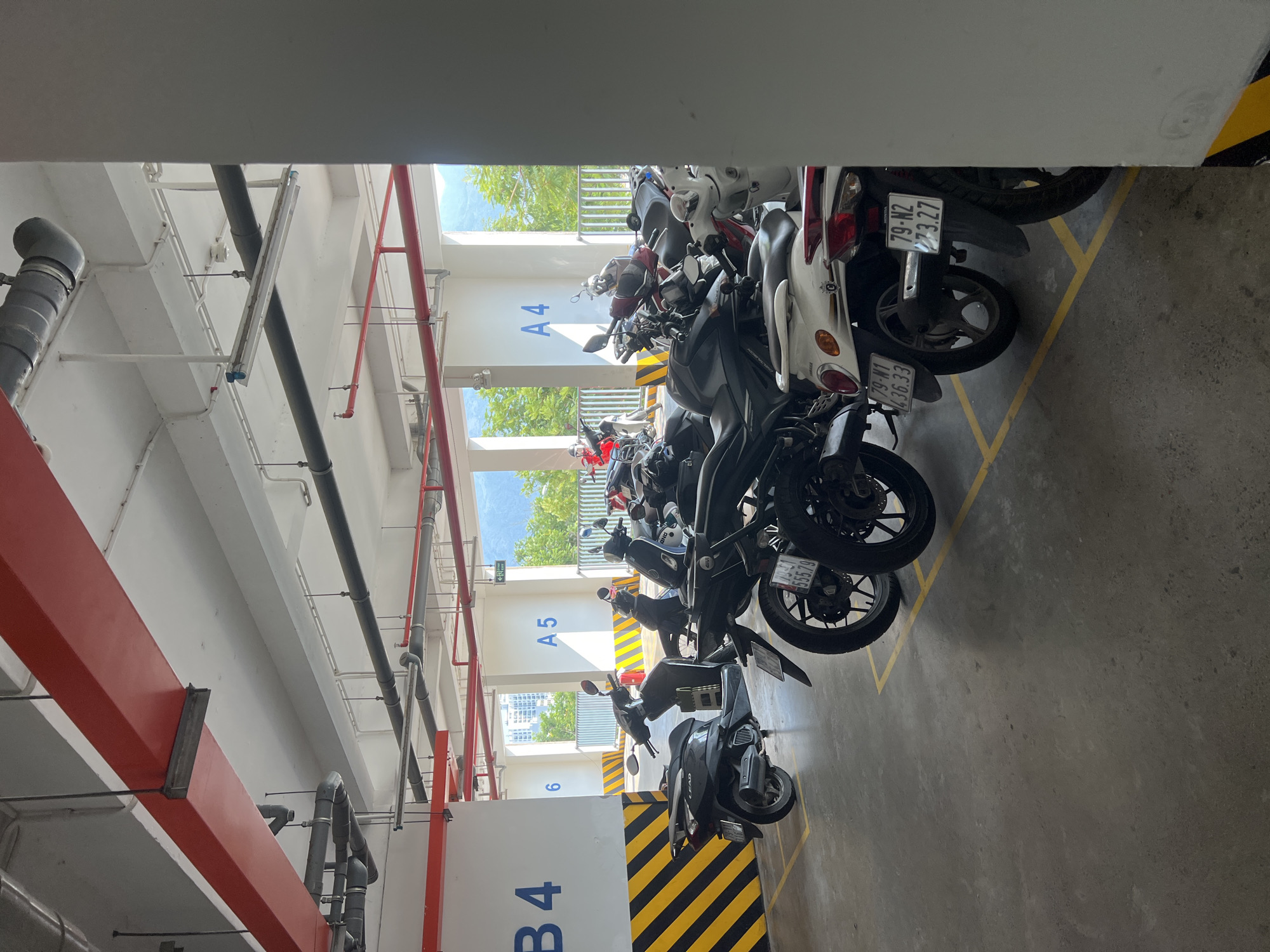 This image depicts a row of parked motorcycles in what appears to be an indoor parking garage. The parking spaces are marked with yellow lines and numbered, with visible signs displaying "A4," "A5," and "B4." The motorcycles vary in color and style. The ceiling of the garage has visible piping and support beams. Through the gaps between the parked motorcycles, trees and buildings can be seen, indicating an exterior view. The location is identified as the Nam Natural Heritage Area Trang, Vietnam.