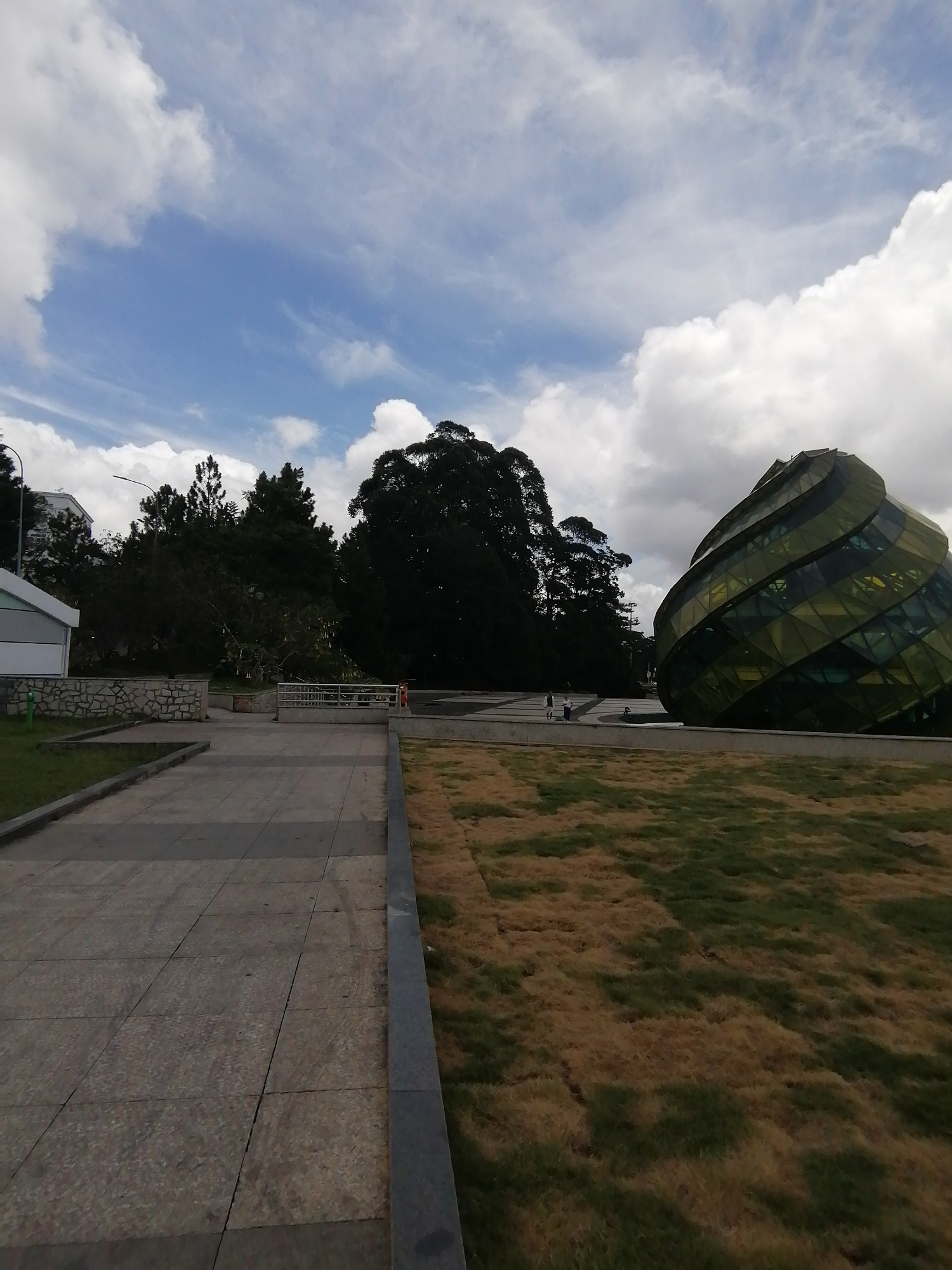 The image depicts a partly cloudy day in Da Lat, Vietnam, with a view of the distinctive glass-domed architecture of the Da Lat Market. A concrete pathway leads from the lower left towards the right, where a large patch of mixed brown and green grass is situated. To the right of the grass, partially framed, is the Da Lat Market's unique dome. The area is surrounded by various trees and foliage, with some trees appearing darker and denser. The sky is partially covered with white and grey clouds. There are no visible interactions taking place, though some people are walking near the Da Lat Market.