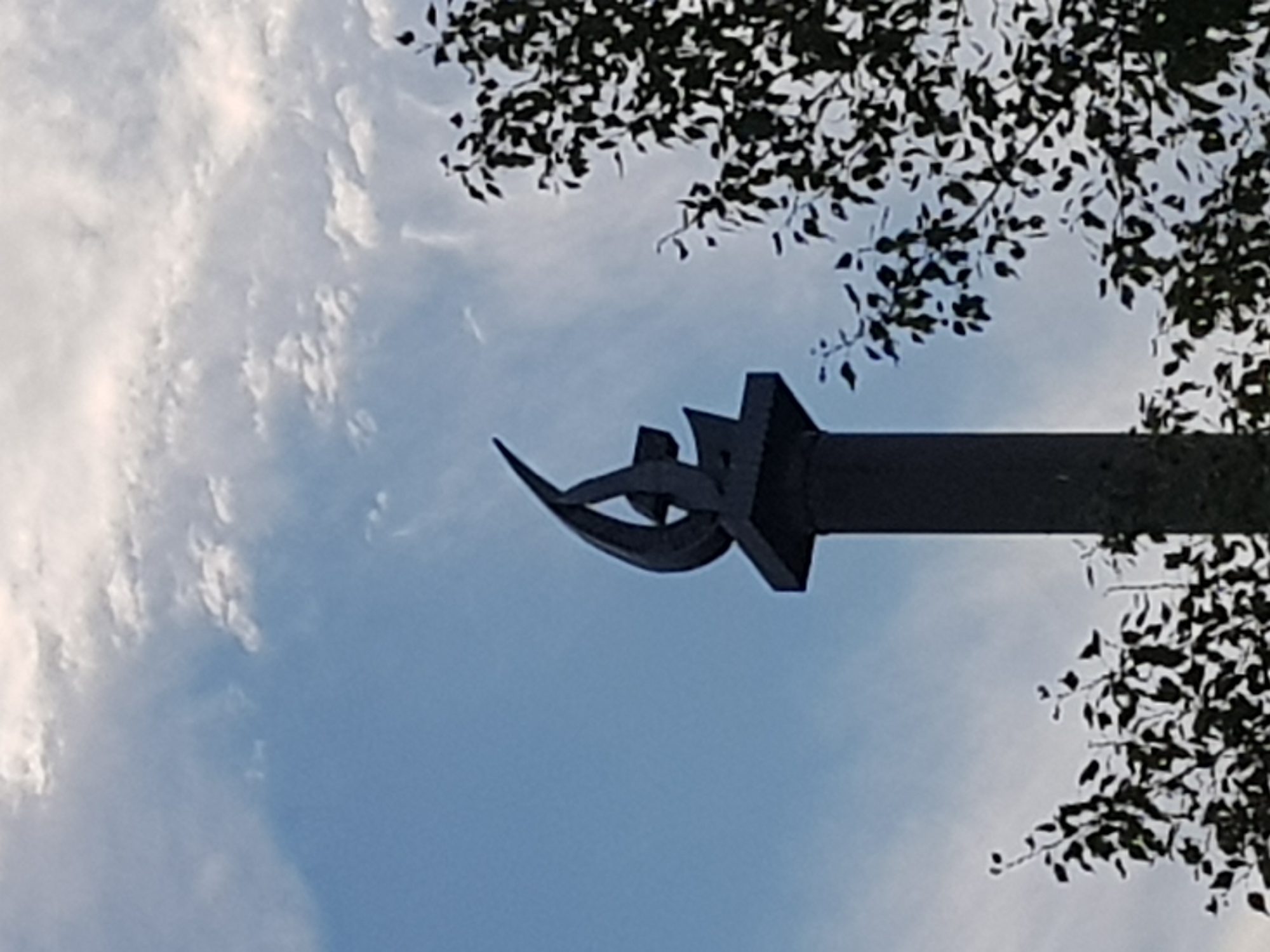 A black, abstract sculpture resembling a flame or stylized bird is perched atop a dark, columnar pedestal. The sculpture features curved, angular elements. The pedestal is positioned outdoors, with a bright blue sky and wispy white clouds visible in the background. To the right and below the pedestal, the dark silhouette of dense foliage from trees is present, with some leaves silhouetted against the sky. The image appears to be taken from a low angle, looking upwards.