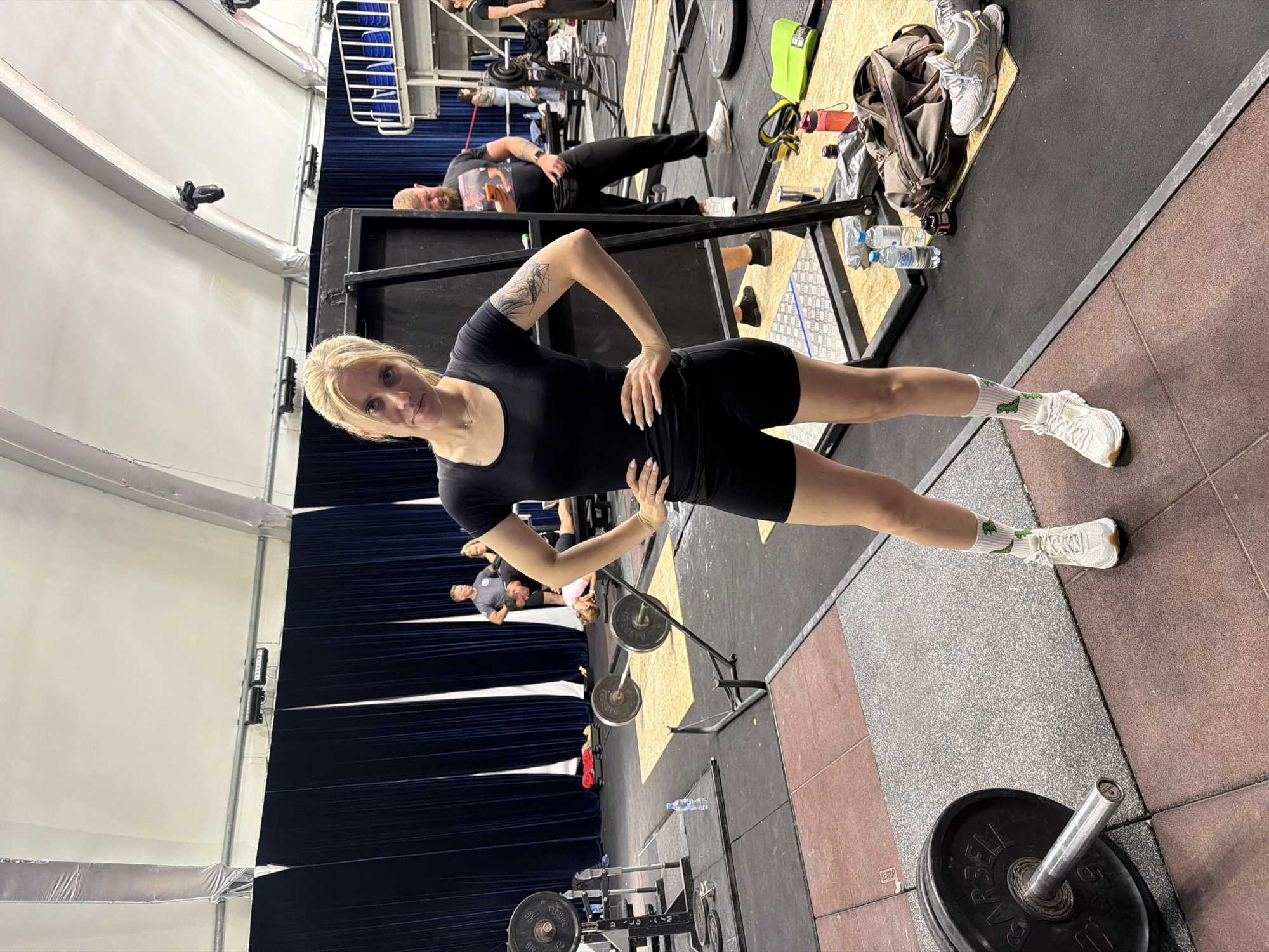 The image shows a woman with blonde hair wearing a black t-shirt and black shorts, standing with her hands on her hips in a gym. She is wearing white sneakers with green dinosaur-patterned socks. The gym appears to be an indoor facility with a large tent-like ceiling. There are weightlifting equipment, including barbells and weight plates, visible in the background. Several other people are in the gym, some appearing to be working out and others observing. The environment is dimly lit with artificial lighting. The image does not contain any information to suggest the location is in Suzdal, Russia.