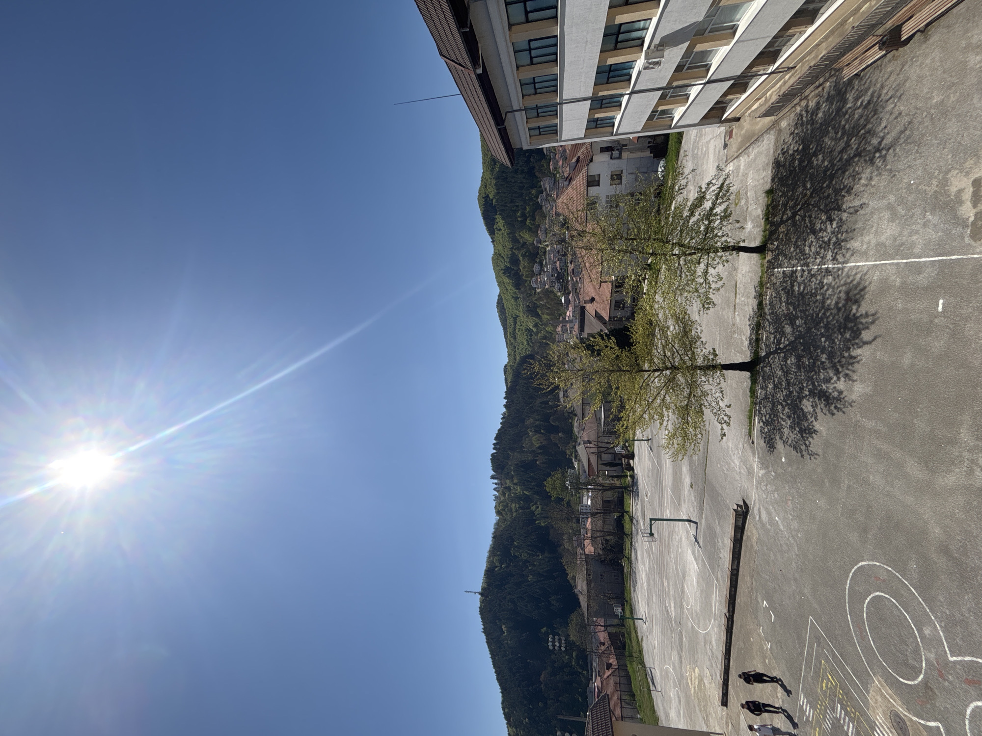 The image, taken in Zlatograd, Bulgaria, presents an outdoor view dominated by a sunlit sports court and a verdant mountain backdrop. The perspective is from an elevated position, looking down onto the scene.
In the foreground, a large asphalt multi-sport court is visible, marked with white lines for basketball, including free-throw lines, a key, and parts of a three-point arc. Other abstract white and yellow markings are also present on the surface. Two basketball hoops with backboards stand on the court. Several trees with fresh green leaves line the left side of the court, casting long, distinct shadows that stretch across the playing area. Three individuals, appearing as silhouettes, stand on the lower right portion of the court, their elongated shadows indicating either an early morning or late afternoon setting. A long wooden bench is placed alongside the court near the adjacent building.
To the right, a multi-story building, likely a school or residential block, features a modern facade with numerous windows and a partially visible dark-tiled roof. A slender lightning rod or antenna extends skyward from the roof.
Behind the court and building, a large, densely forested mountain rises, covered in lush green trees. Houses with characteristic reddish-brown tiled roofs are nestled on the lower slopes of the mountain, blending into the natural landscape. The sky above is a brilliant, cloudless blue, with the bright sun positioned in the upper left, causing a noticeable lens flare. The clear sky and strong shadows indicate a bright, sunny day. No legible text is visible in the image.