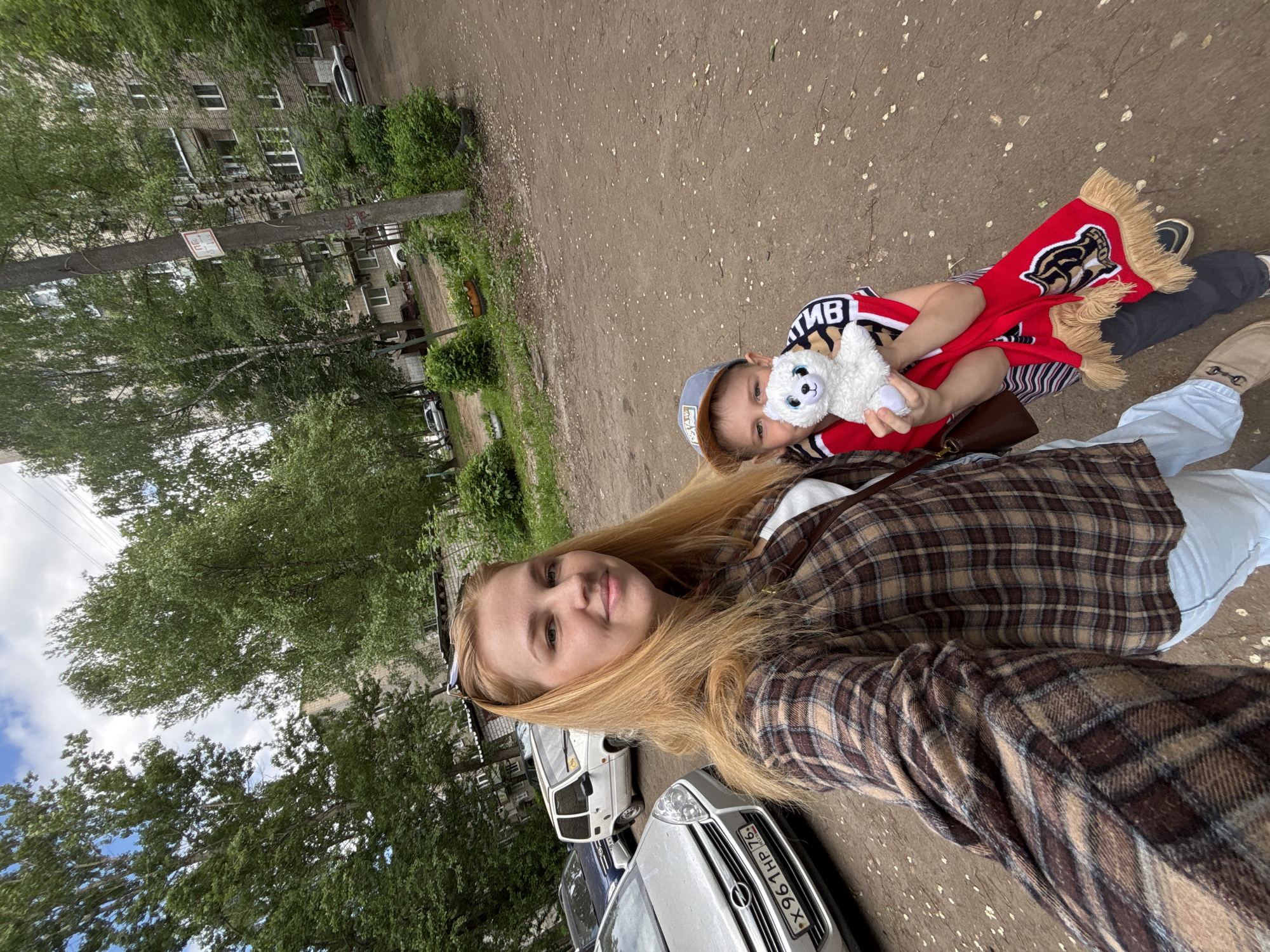 A selfie-style photograph shows a woman with long blonde hair, wearing a plaid shirt and light blue jeans. She is holding a young child wearing a striped shirt, cap, and a red scarf with a bear logo, who is holding a stuffed polar bear. The photo is taken outdoors on a paved area with trees and buildings in the background. Cars are visible on the side. The license plate on a silver Opel is X961HP 76, indicating the Yaroslavl Oblast of Russia. Based on the setting, the photograph was likely taken in the city of Rusanovo, Russia.