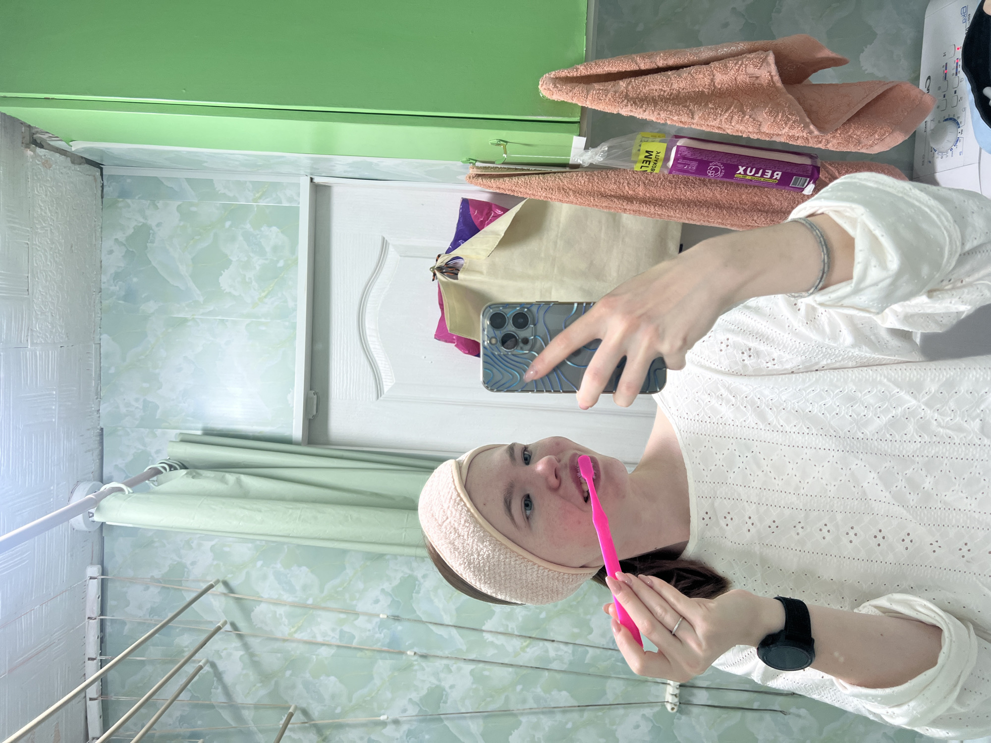 A person is shown in a bathroom, taking a selfie while brushing their teeth. The person is wearing a white top and a headband. The background includes bathroom fixtures such as a drying rack, shower curtain, washing machine, towels, and various toiletries. The location is Chaikovsky, Russia.