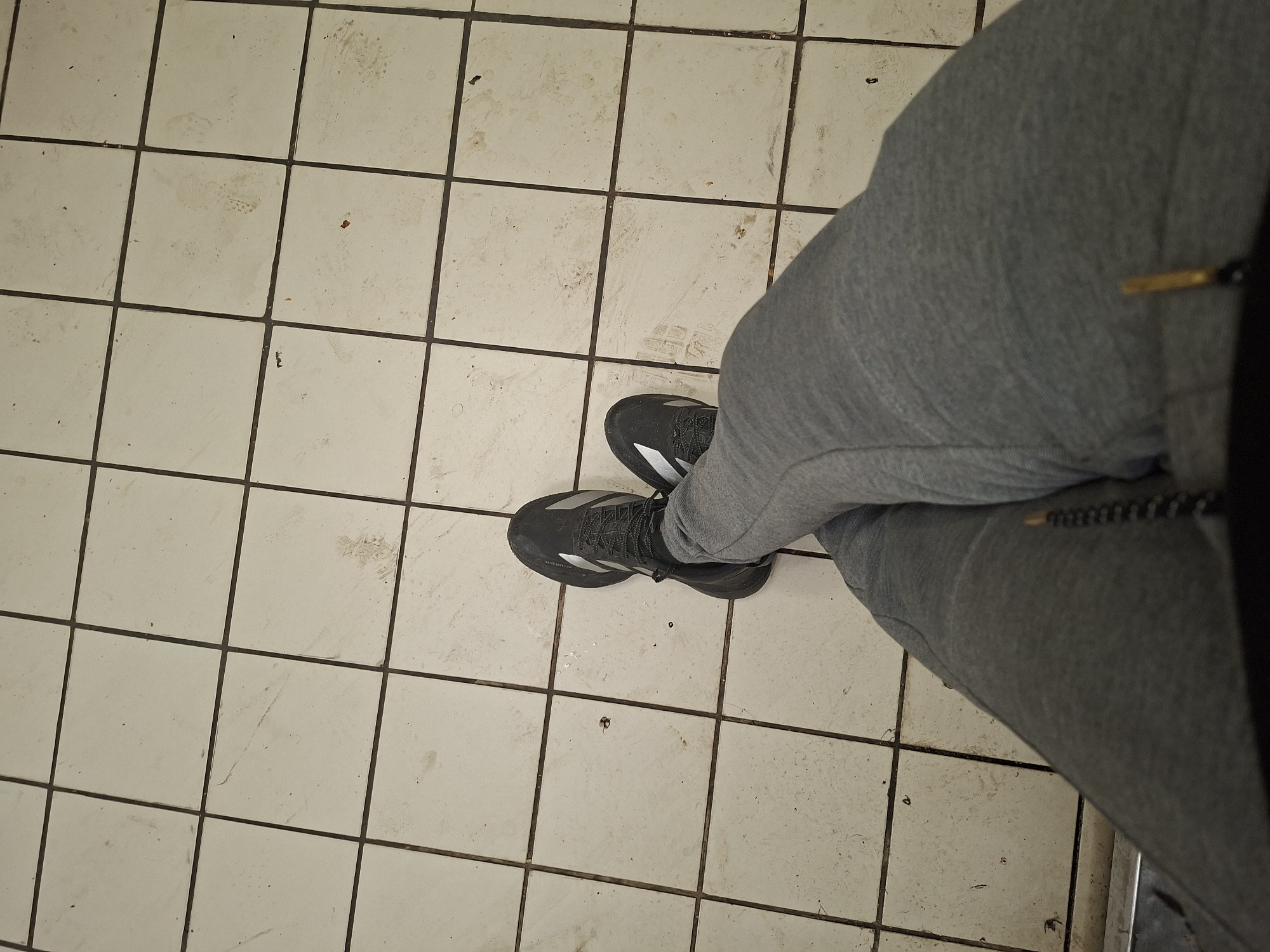 A first-person perspective shot shows a person's legs and feet, clad in grey sweatpants and black athletic shoes with white accents, resting on a tiled floor. The floor is composed of large, off-white square tiles with dark grout lines. The tiles appear somewhat worn and dirty, with scuff marks and smudges visible. The lighting is dim, suggesting an indoor environment, possibly a locker room, hallway, or a similar utilitarian space. The background is out of focus, but the grid pattern of the tiles dominates the scene. There is no discernible indication of the location being specifically within the city of Nicosia, Cyprus. The overall impression is one of a casual, perhaps mundane, moment captured.