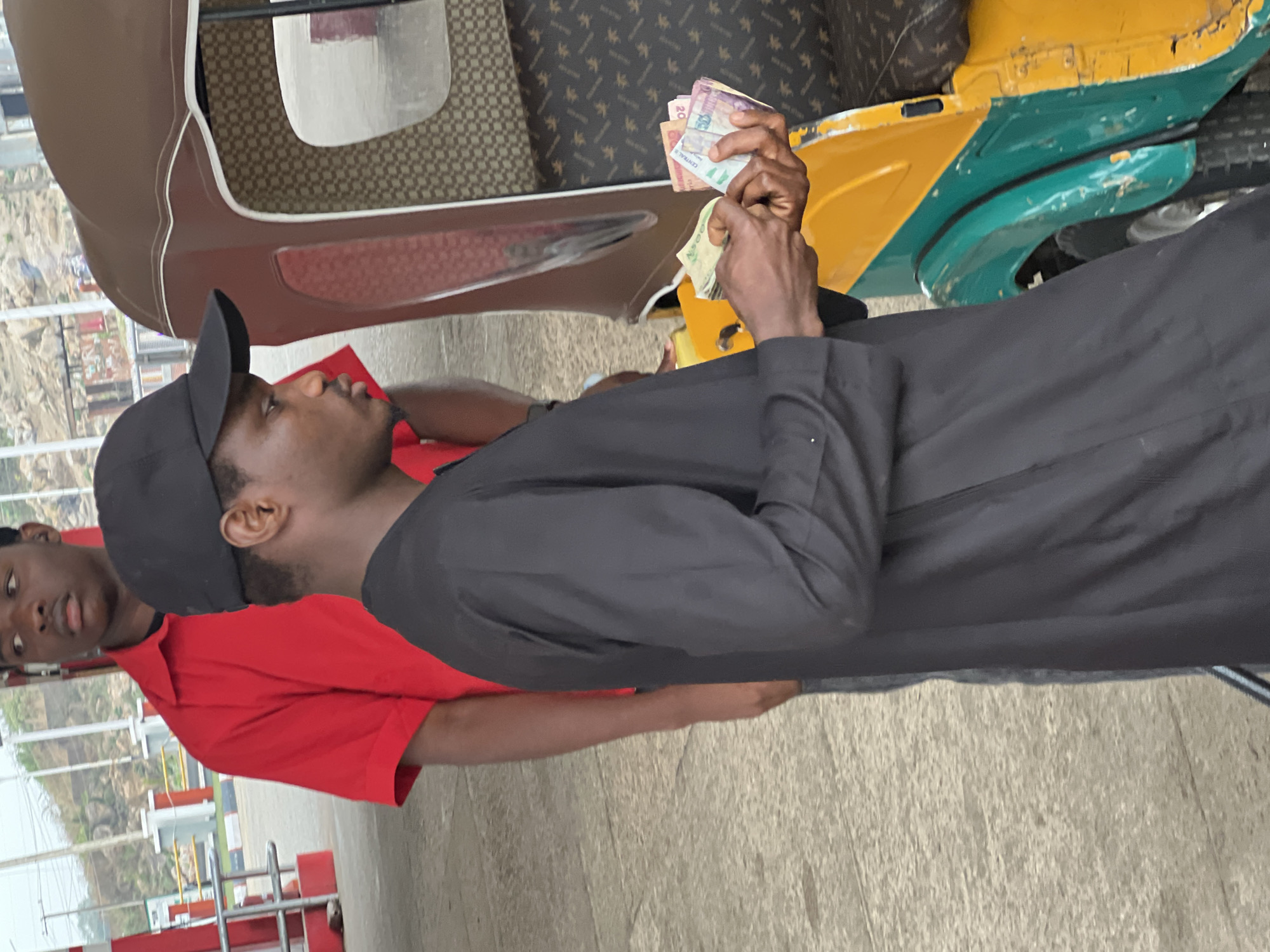 A ground-level, medium shot taken outdoors shows two young men and a yellow and green auto rickshaw, also known as a Keke Napep. The man in the foreground is facing to the left and counting a wad of Nigerian naira bills. He is wearing a black cap, a black long-sleeved shirt, and gray pants. Behind him to the left is a second man wearing a red shirt. He is looking at the man counting money. The auto rickshaw is partially visible to the right, with brown and patterned upholstery in the passenger area.
The location appears to be in the city of Jos, Nigeria. The background shows buildings with green landscaping and a fence. In the extreme left is a red metal fence or gate with a vertical stripe pattern. The ground is a light-colored concrete slab.