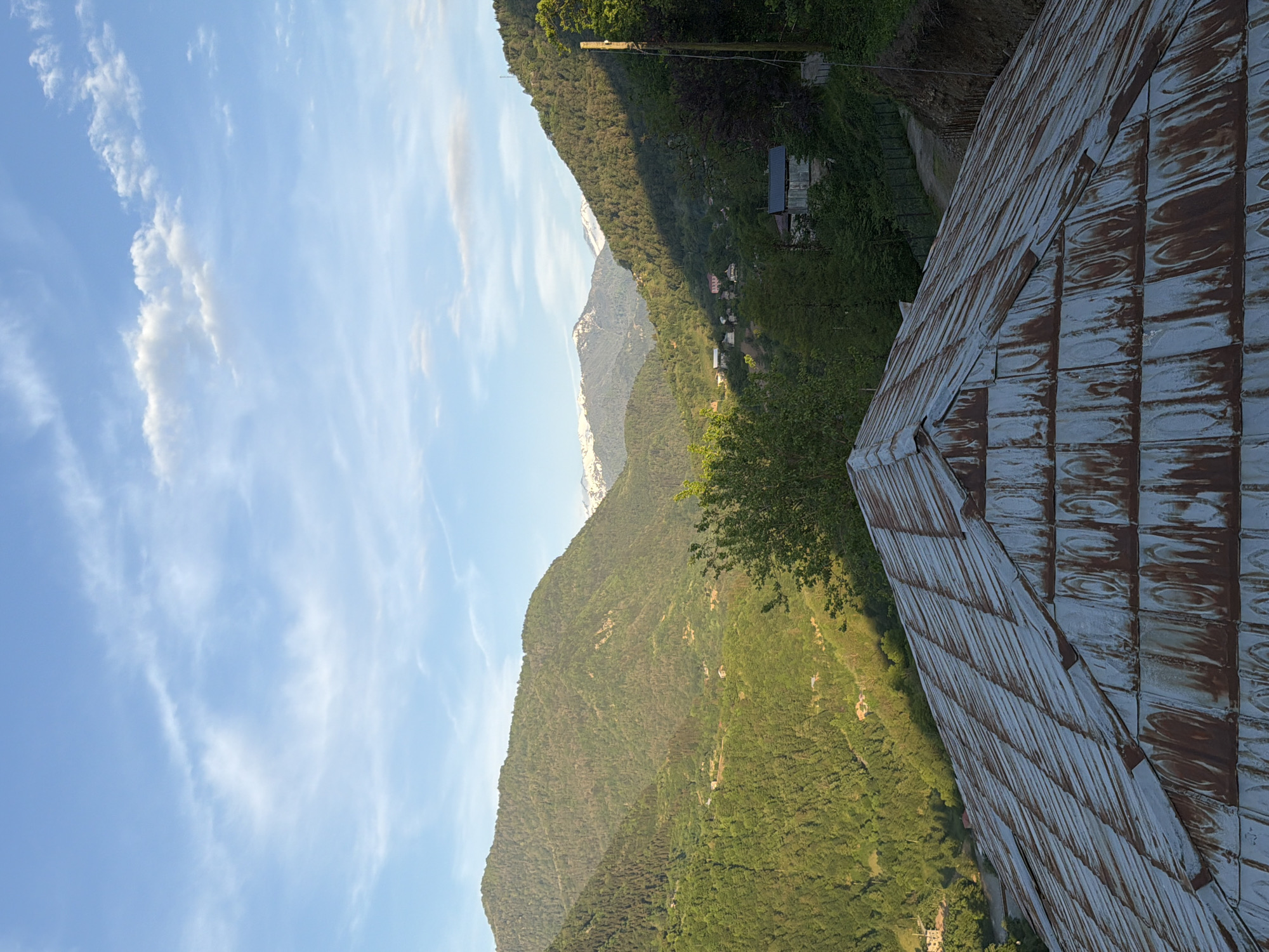 The image shows a landscape near Kveda Chkhutuneti, Georgia. The shot is angled, presenting a view of mountain ranges with a clear blue sky interspersed with light clouds. The mountains are covered with green vegetation, and a second mountain range in the distance appears to have snow-capped peaks. In the foreground, a corrugated metal roof is visible, occupying a significant portion of the right side of the frame. The structure associated with the roof is not entirely in view.