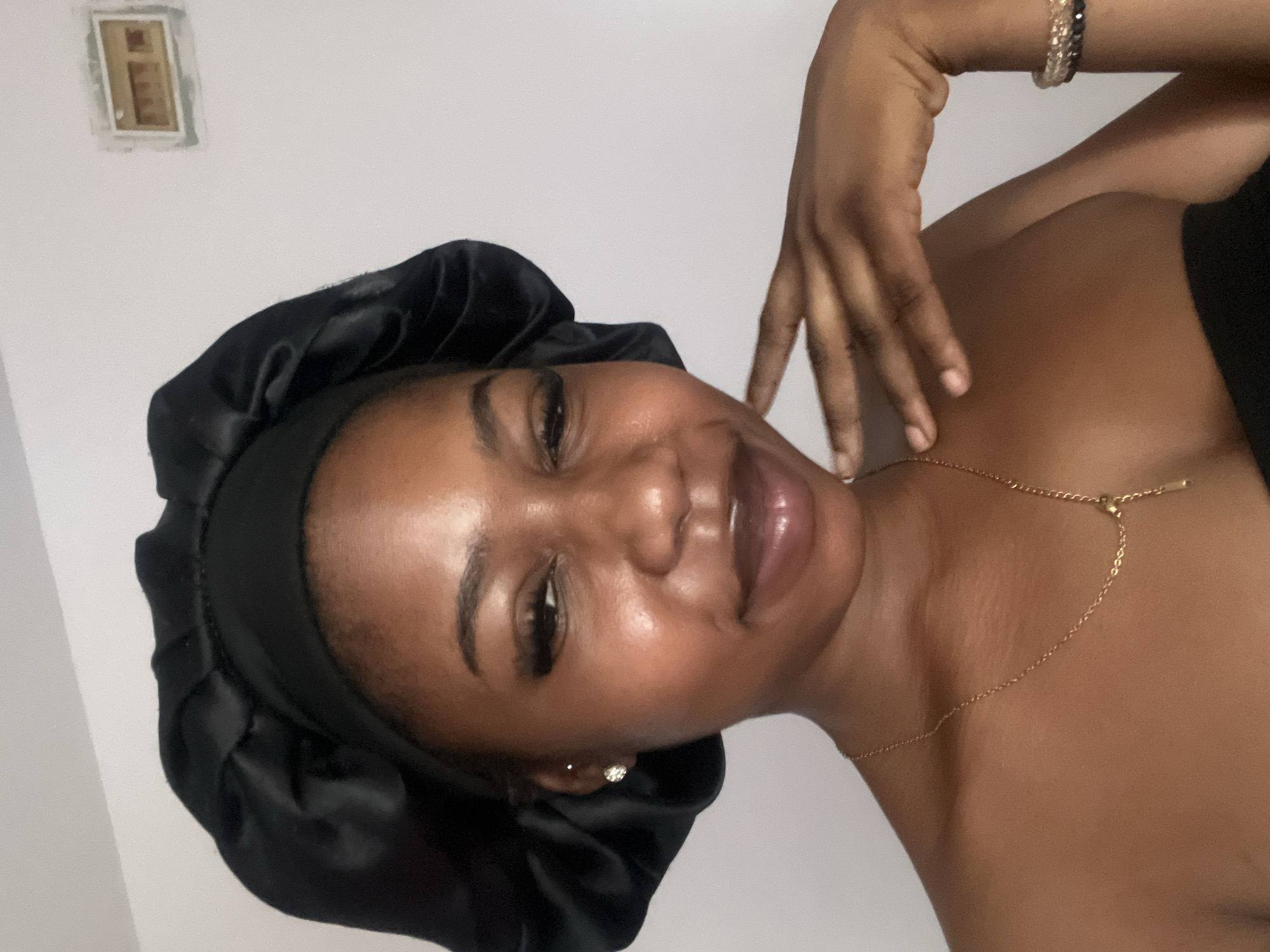 A young Black woman wearing a black satin bonnet and a gold necklace is pictured in a close-up, head-and-shoulders shot. She is looking towards the viewer with her eyes partially closed and a slight smile on her lips. Her left hand is raised, with her fingers resting near her face. She is wearing a small stud earring in her left ear and a bracelet on her right wrist. The background is a plain, light-colored wall. There is a light switch cover plate visible on the wall to the upper right of the woman. The image appears to be taken indoors, possibly in a residential setting in Jos, Nigeria.