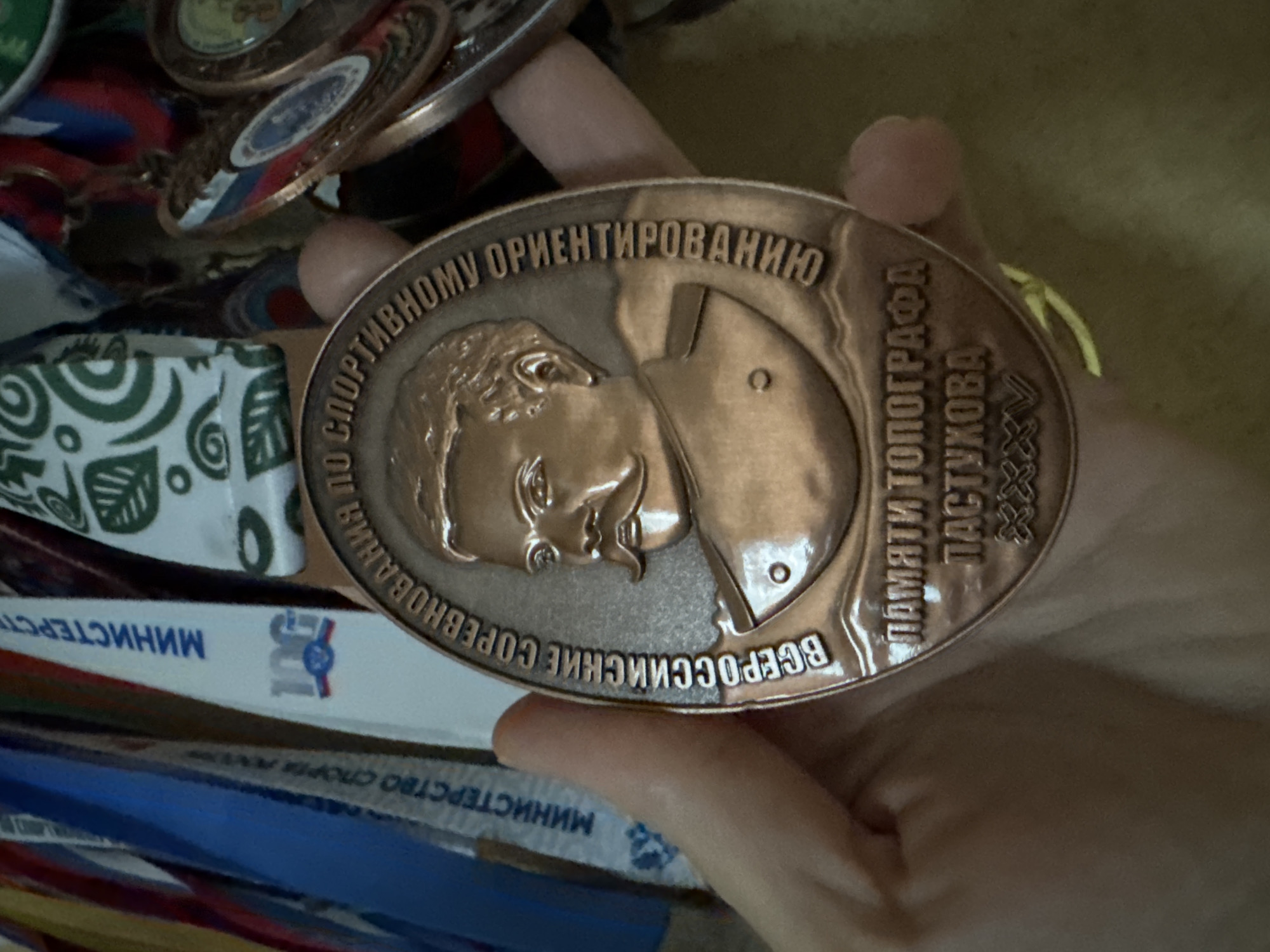 In this close-up shot, a person's hand is holding a round, copper-colored medal. The medal features an embossed profile of a man with a mustache, wearing a uniform. The man's face is depicted in profile, facing left, with his hair and facial features clearly defined. Below the portrait, text is inscribed in what appears to be Russian. The surrounding area shows the edges of other medals and ribbons, suggesting they are part of a collection or award display. Some of these ribbons are blue, white, and red, colors often associated with national flags. In the background, parts of other medals with intricate designs and colorful ribbons are visible. The overall lighting is somewhat dim, highlighting the metallic sheen of the central medal.