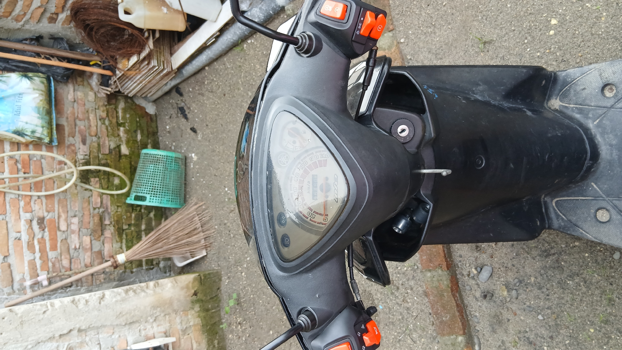 This is a close-up, eye-level, still photograph of the front handlebars and dashboard of a black scooter parked outdoors. The scooter's dashboard features a speedometer and tachometer with a digital odometer displaying "051604". Orange and black control buttons are visible on the left and right handlebars. In the background, a brick wall is present, with various items leaning against it, including a broom, a green plastic basket, coiled wire, and a plastic jug. The ground appears to be a mix of dirt and gravel. The scene is located in Klambir Lima Kebun, Indonesia.