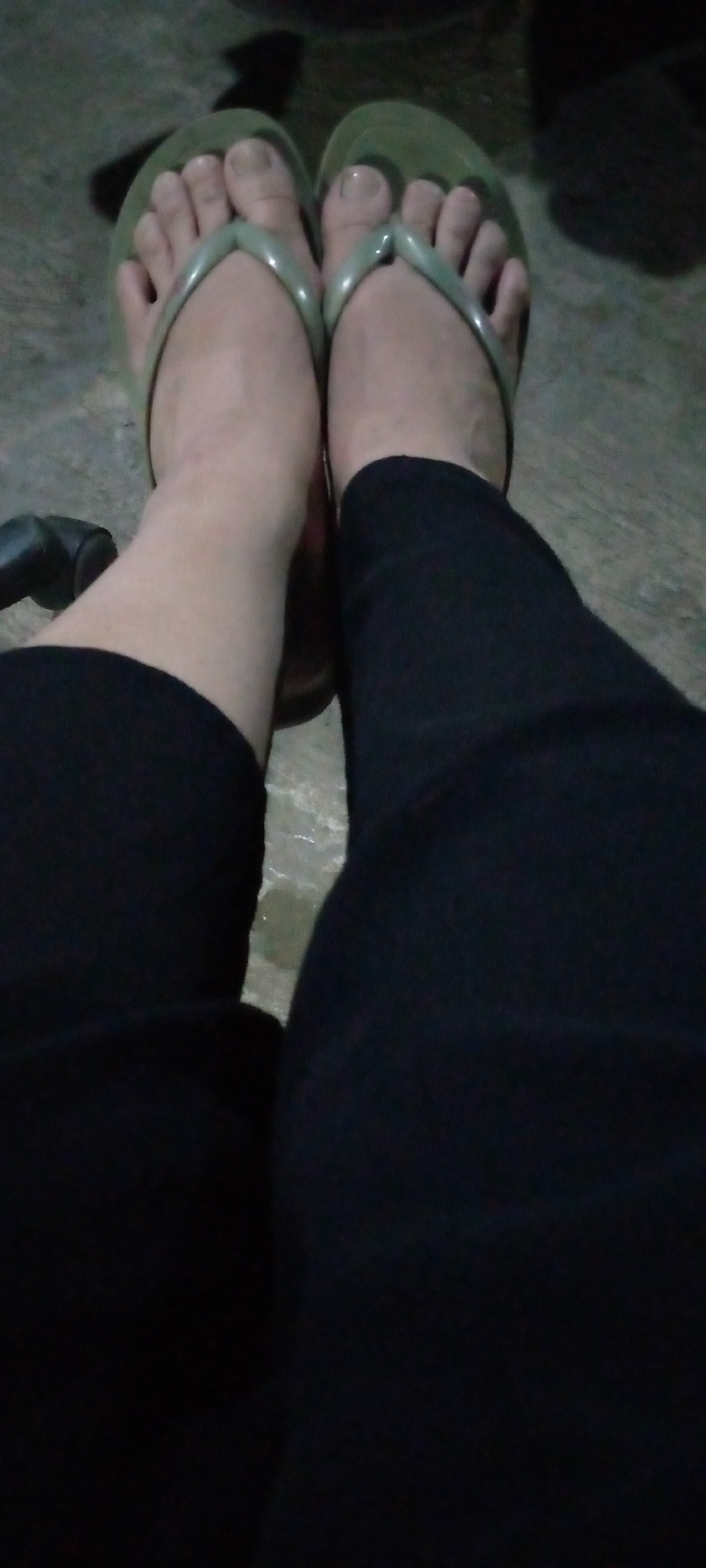 The image shows a close-up of a person's feet wearing green flip-flops and black leggings. The feet are resting on a concrete surface, which appears to be outdoors. The lighting is dim, suggesting it might be evening or a poorly lit area. There is a dark, amorphous shadow in the background, and a dark object is partially visible on the left side of the frame. No specific location in Sumberwono, Indonesia can be identified from the visual information provided. The image focuses solely on the lower extremities of an individual.