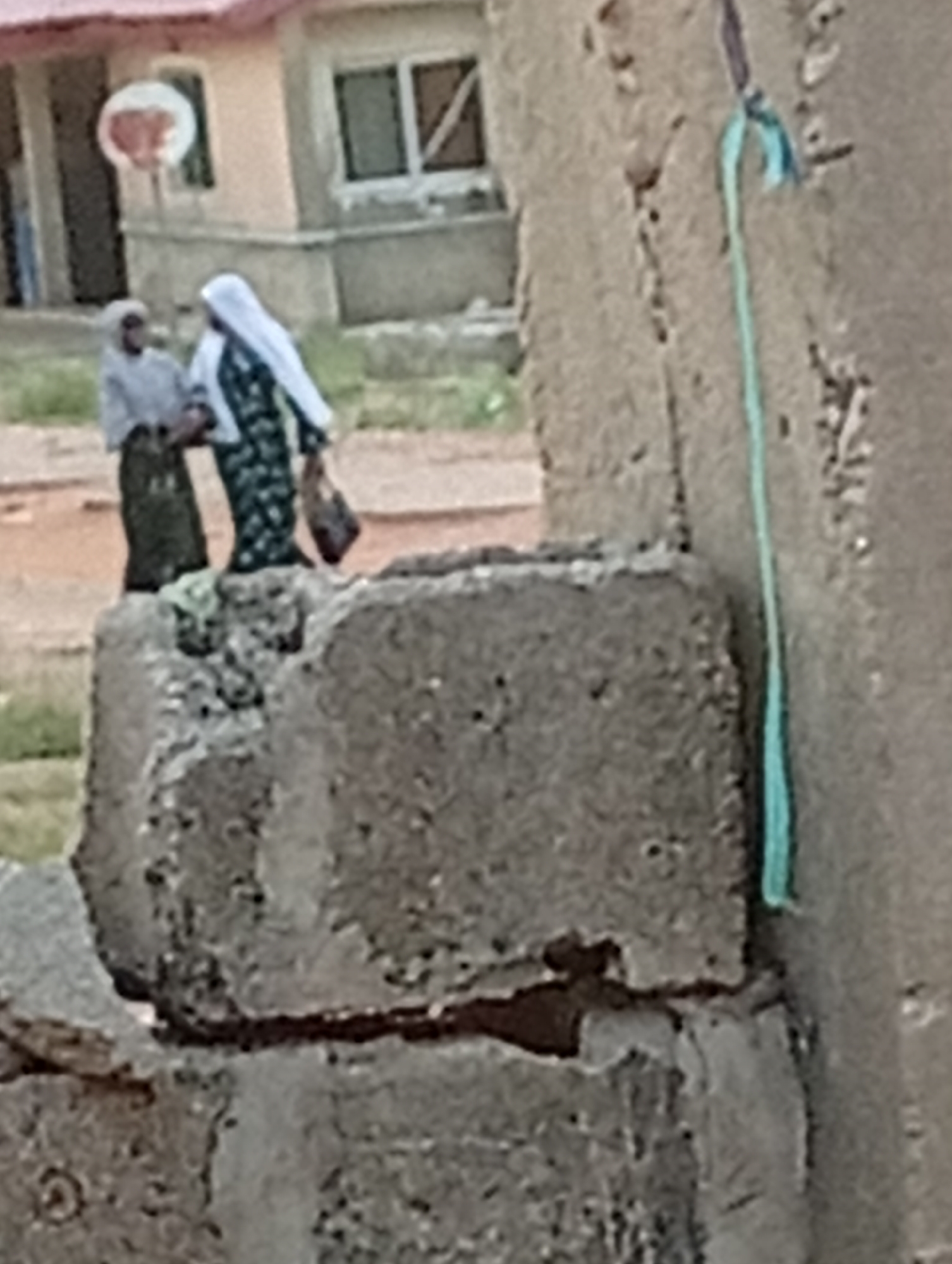 In Adankolo, Nigeria, two people stand behind a concrete structure. One person wears a light grey head covering, a light grey top, and a dark skirt. The other person wears a white head covering and a dark patterned dress and carries a bag. A building and a basketball hoop are visible in the background. A green rope hangs on the right side of the concrete structure.