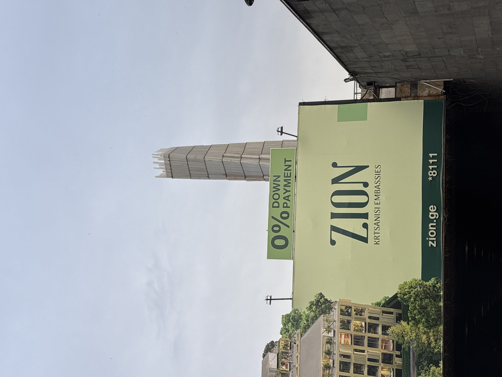 The image, rotated correctly, depicts a daytime urban scene featuring a large billboard prominently in the foreground, with a tall building under construction behind it. The sky is a bright, light blue with minimal cloud cover.
The billboard, primarily light green, advertises a real estate development. The main text, "ZION," is displayed in large, dark green letters, with "KRTSANISI EMBASSIES" written beneath it in a smaller font. A significant offer, "0% DOWN PAYMENT," is highlighted in a lighter green box on the left side of the billboard. At the bottom right, the website "zion.ge" and a phone number "*8111" are visible on a darker green strip. The billboard also includes an artistic rendering of modern buildings integrated with lush greenery and trees.
Behind and to the left of the billboard, a very tall, slender structure rises against the sky. It appears to be a building in an advanced stage of construction, possibly enveloped in scaffolding or a protective wrap, giving it a textured, rectangular profile. A dark, concrete-like overhead structure, likely part of an underpass or bridge, partially frames the scene, visible at the top and bottom right edges of the image. A small pole with a fixture extends upwards behind the billboard.
The visible text, especially "KRTSANISI EMBASSIES" and the '.ge' domain, strongly indicates the location is Tbilisi, Georgia, specifically referencing the Krtsanisi district. No human subjects or specific activities are depicted; the scene is a static urban snapshot.