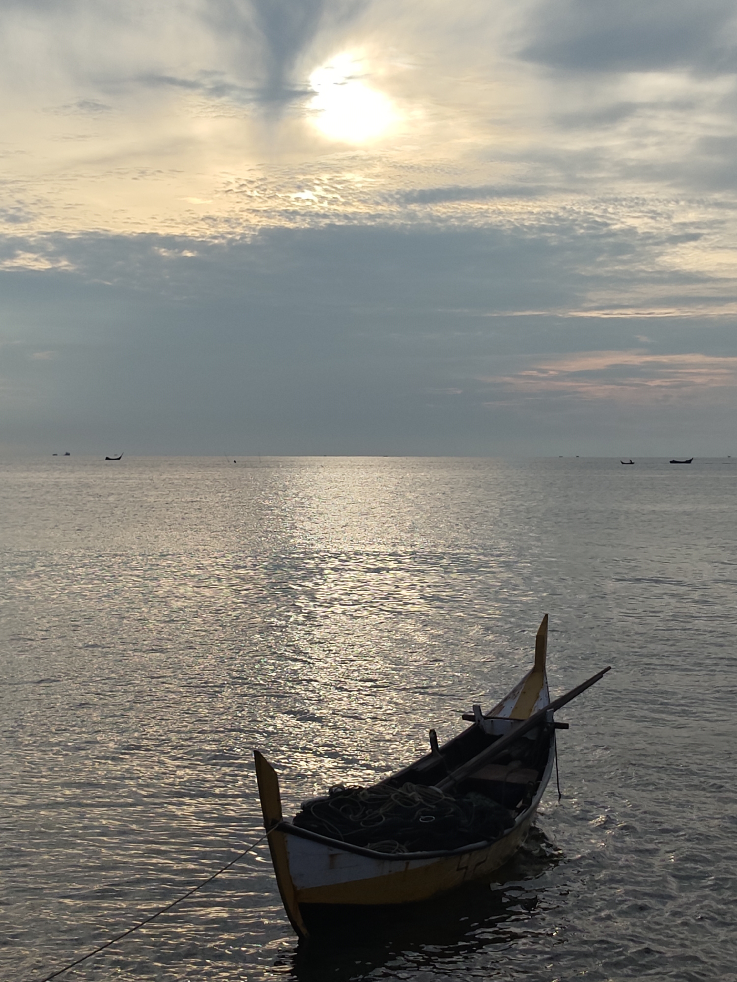 A fishing boat, yellow and white, floats on calm water under a cloudy sky with the sun peeking through. The sun creates a shimmering reflection on the water's surface. In the distance, other boats can be seen on the horizon, suggesting it is a coastal area. The scene is likely set in Lhokseumawe, Indonesia, a city known for its coastline and fishing activities. The image captures a peaceful, serene moment, possibly at sunrise or sunset.