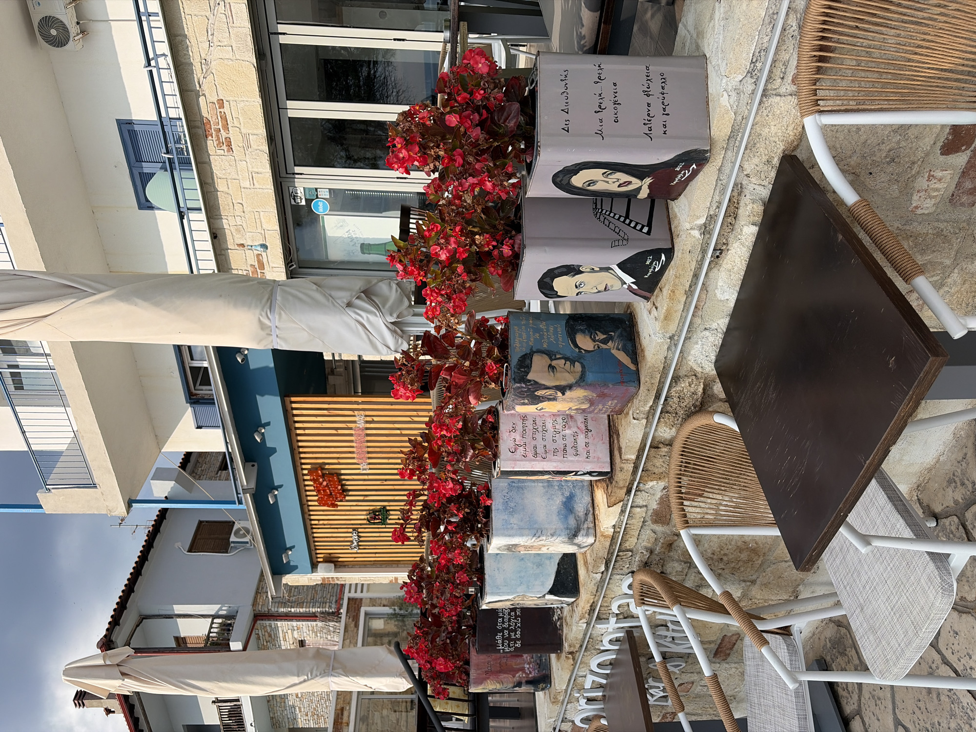 In the city of Kassandra Municipal Unit, Greece, an outdoor seating area features a row of decorative planters filled with vibrant red flowers. The planters are square and painted with portraits of individuals and Greek text. These planters are placed on a stone ledge in front of a building with a blue awning and wooden slatted walls. Two tables with dark wooden tops and white chairs with woven seats are visible in the foreground. The chairs have decorative rope detailing on their arms. Behind the planters, the building has large glass windows, and a covered outdoor umbrella stands to the left. The sky is partly cloudy.
