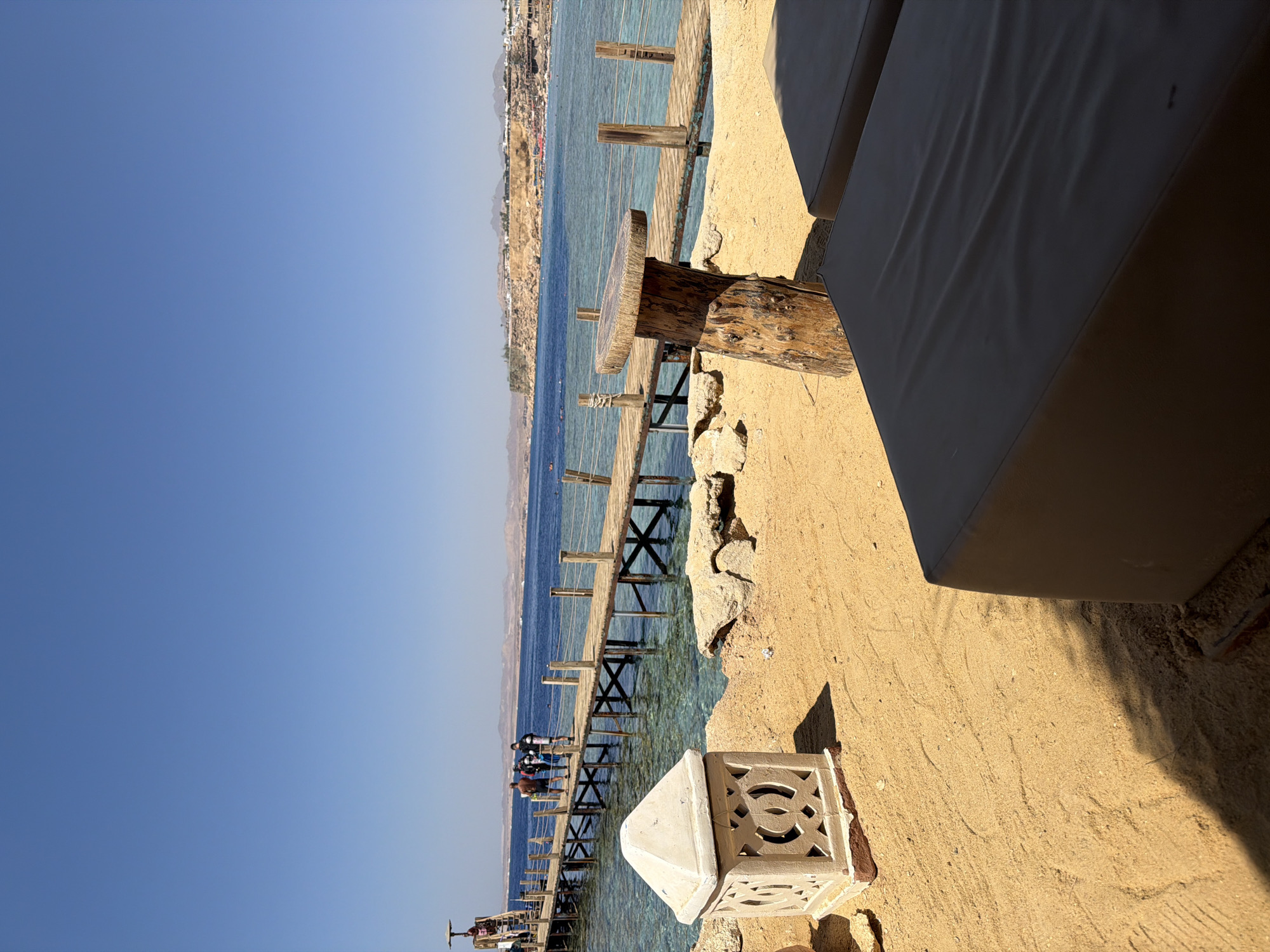 The image depicts a serene beach scene in Sharm El Sheikh, Egypt, under a clear blue sky. In the foreground, two empty dark-colored lounge chairs rest on a sandy beach. To the left of the chairs, a decorative, geometric stone lantern sits on the sand, casting a shadow. A rustic wooden table made from a tree trunk and a thick slice of wood stands between the lounge chairs and a wooden pier.
The pier extends over the turquoise water, supported by wooden pilings. Several people, including individuals wearing scuba gear, are visible walking along the pier. The water is clear, revealing the seabed in some areas. In the background, the coastline of Sharm El Sheikh is visible, characterized by rocky hills and scattered buildings, suggesting a resort area. The overall atmosphere is tranquil and inviting, typical of a beach destination.