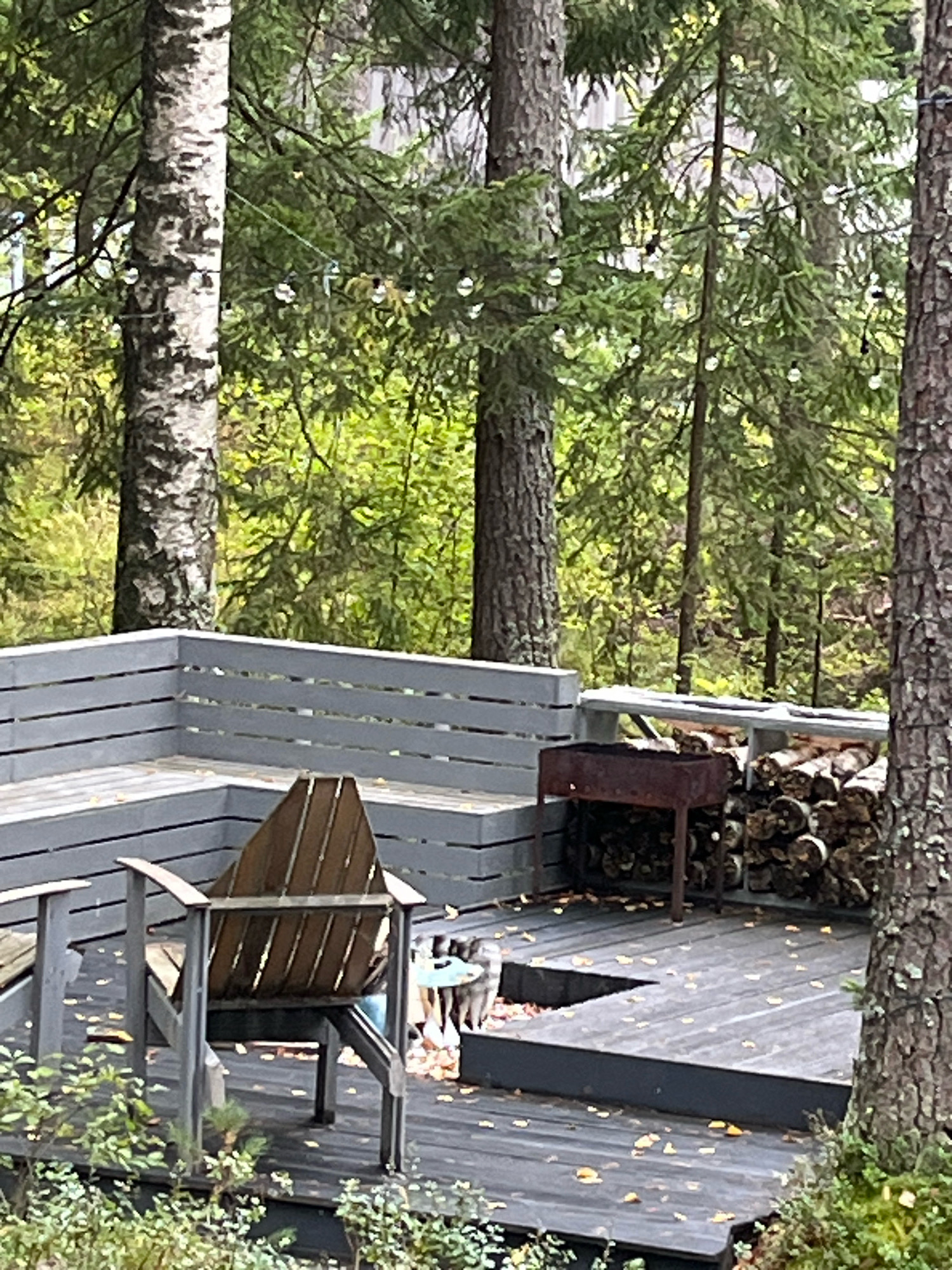 A wooden deck with a grey railing is set amongst trees. On the deck, there is a wooden chair, a charcoal grill, and a pile of chopped firewood. The deck appears to be in a wooded area.