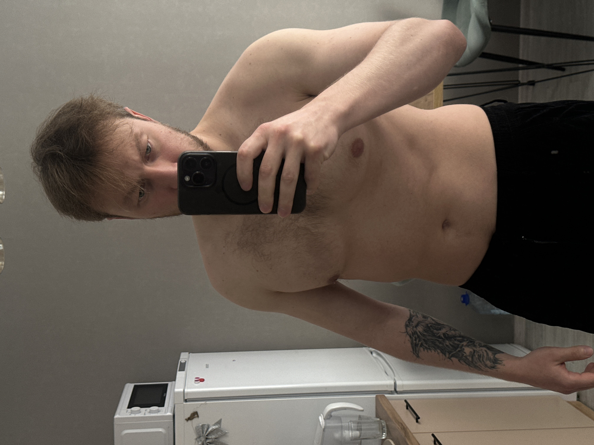 A shirtless man is holding a smartphone in his right hand, presumably taking a selfie. He is wearing black shorts. He has light brown hair and a beard. His left arm has a tattoo. Behind him is a white refrigerator, a microwave, and a kitchen counter. The environment suggests a domestic interior. There is no evidence to support a location in Sochi, Russia.