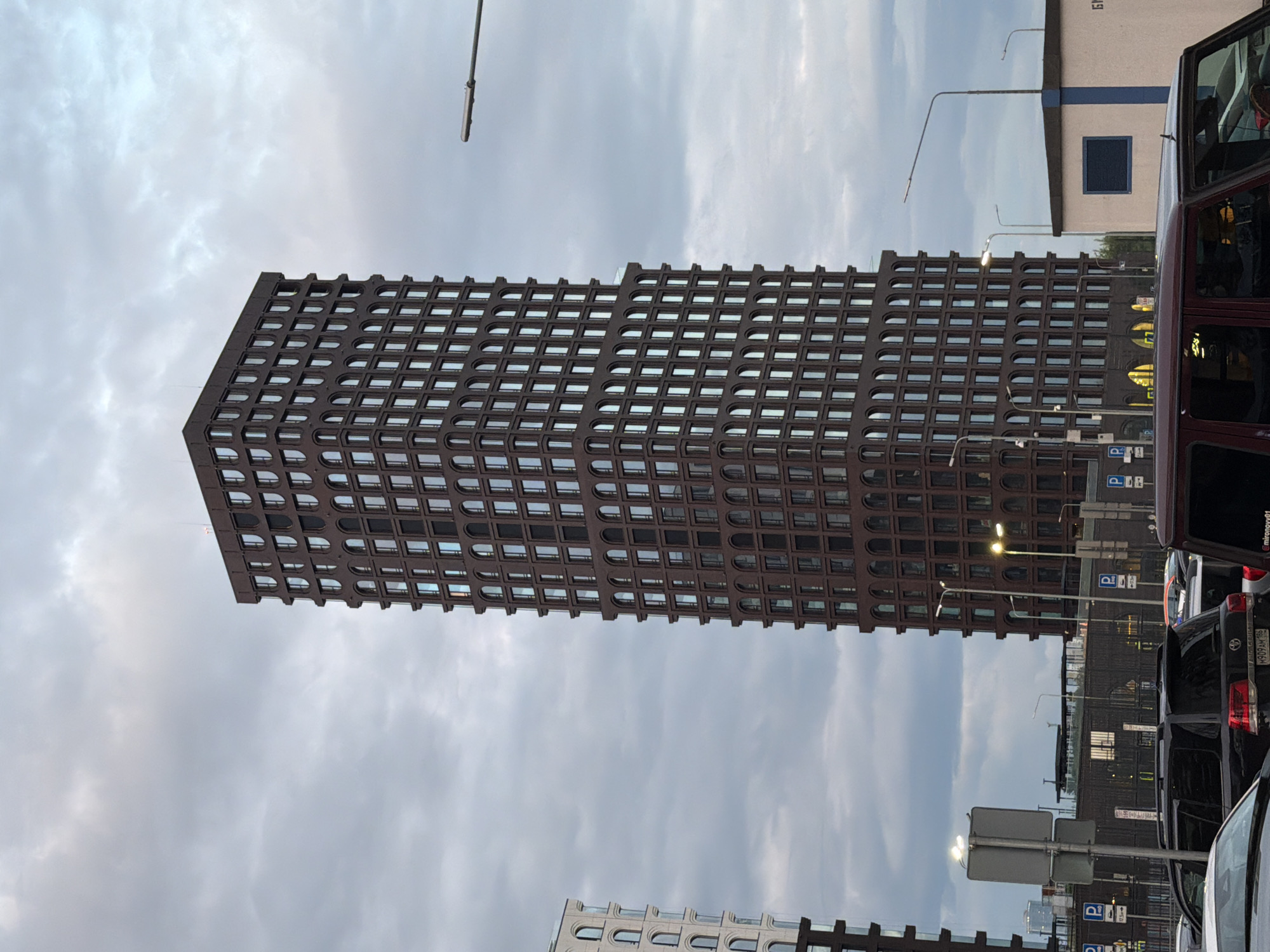 This media file captures a street scene in Kazan, Russia, with a prominent, tall, dark brown building with a grid of windows dominating the left side of the frame. The building appears to be modern with repeating arched window designs. In the background, other buildings are visible, including a white and blue structure. The sky is cloudy and overcast, suggesting a possibly cool or temperate day. The time of day is unclear due to the lighting conditions. Several vehicles, including dark-colored SUVs and a white car, are parked or driving on the street, indicating urban activity. One of the vehicles has a license plate with Russian characters. Signs with parking symbols (P) are visible, reinforcing the urban setting. The overall mood of the scene is neutral, focusing on the architectural and urban elements. There is no visible text in the primary subject building, but one of the cars has a license plate with Russian text.