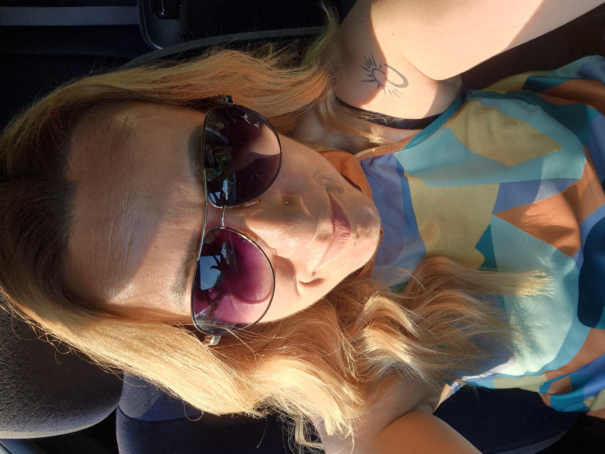 A woman with blonde hair and sunglasses is seen from the chest up, taking a selfie in what appears to be a car. Sunlight is shining on her face, creating shadows. She is wearing a colorful, abstract-patterned tank top. A small tattoo of a sun and moon is visible on her upper arm. The background is dark and out of focus, with a hint of a car seat.