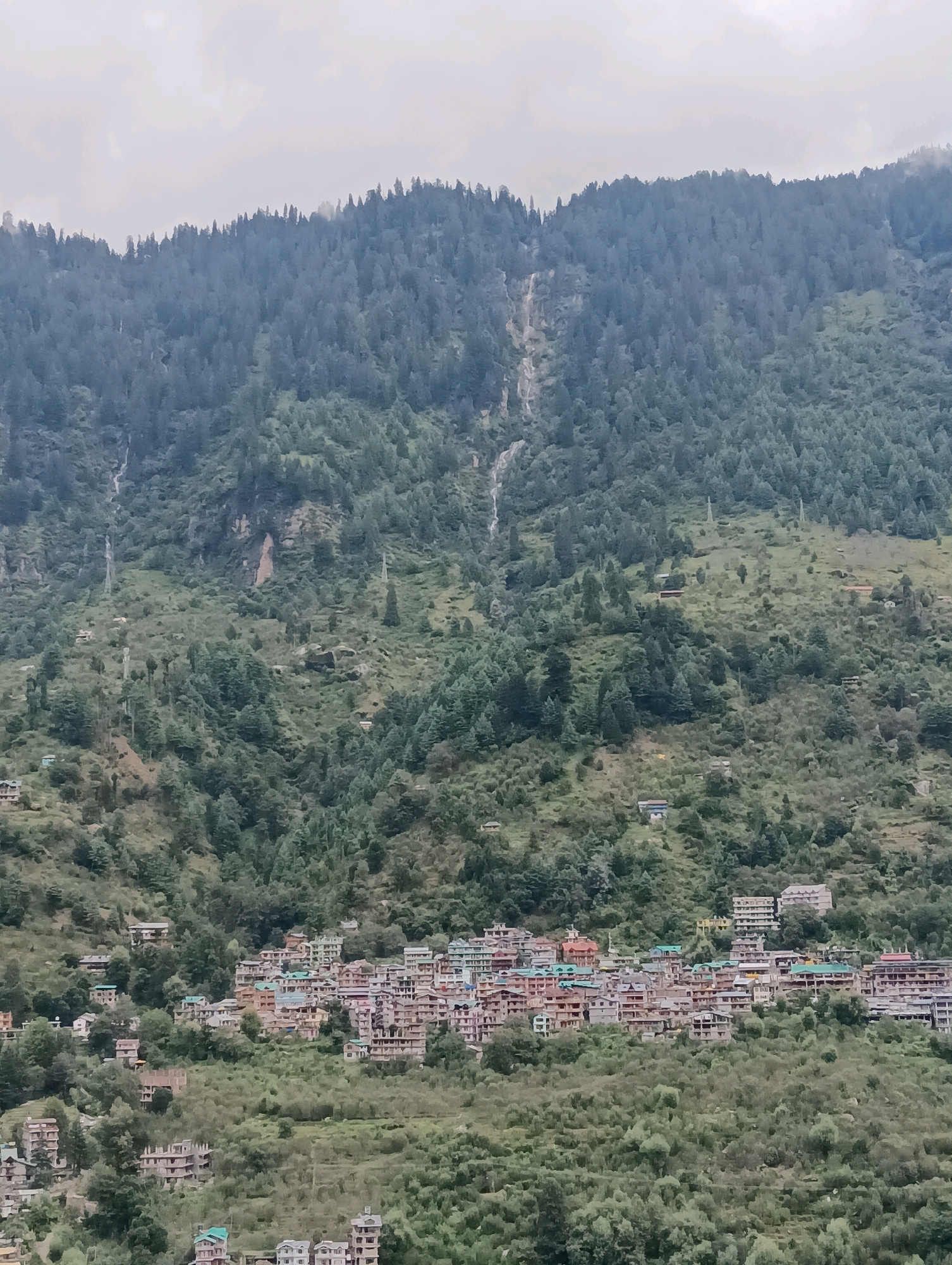 The image depicts a village nestled on a mountainside in Vashisht, India. The village is characterized by a cluster of buildings with colorful roofs, predominantly red and green, built on a steep incline. The architecture features multiple stories and balconies, suggesting a dense settlement. The surrounding environment is dominated by a dense forest covering the mountain, with visible waterfalls cascading down the slopes. In the foreground and scattered across the lower slopes are more modern buildings, some appearing to be under construction. The sky is overcast, casting a soft, diffused light over the scene. The overall impression is of a remote, mountainous community integrated with its natural surroundings.