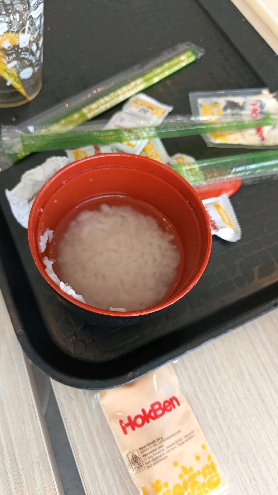 The image displays a tray with food items. In the center of the tray is a red bowl filled with white rice and water. To the left of the bowl is a clear plastic cup with some liquid and yellow and white designs. To the right of the bowl are several packets and green plastic-wrapped chopsticks. The packets appear to be condiment packets, with some displaying text that reads "HokBen". The tray is black and appears to be made of plastic. The background shows a wooden surface, likely a table. The scene is indoors, possibly at a food establishment. Given the presence of "HokBen" branding, this image might be from a HokBen restaurant or outlet in Karawang, Indonesia, a city known for its industrial sector and growing commercial areas.