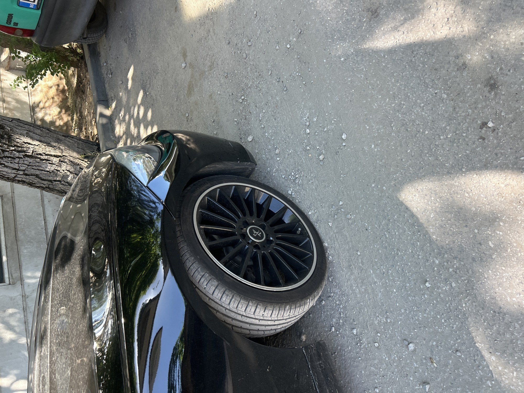 A close-up, high-angle, outdoor shot shows the front quarter panel and wheel of a black car. The car is parked on a light grey gravel surface. The car's tire is black with a silver rim, and the car's body is glossy and reflective, showing hints of trees and sunlight. To the right of the black car, a small portion of the rear of a teal-colored car is visible. The teal car has a license plate with blue characters and a green background. Behind the cars, a rough, textured concrete wall runs horizontally, with a large tree trunk and branches casting shadows on the ground. The area appears to be a street or parking lot in an urban environment.