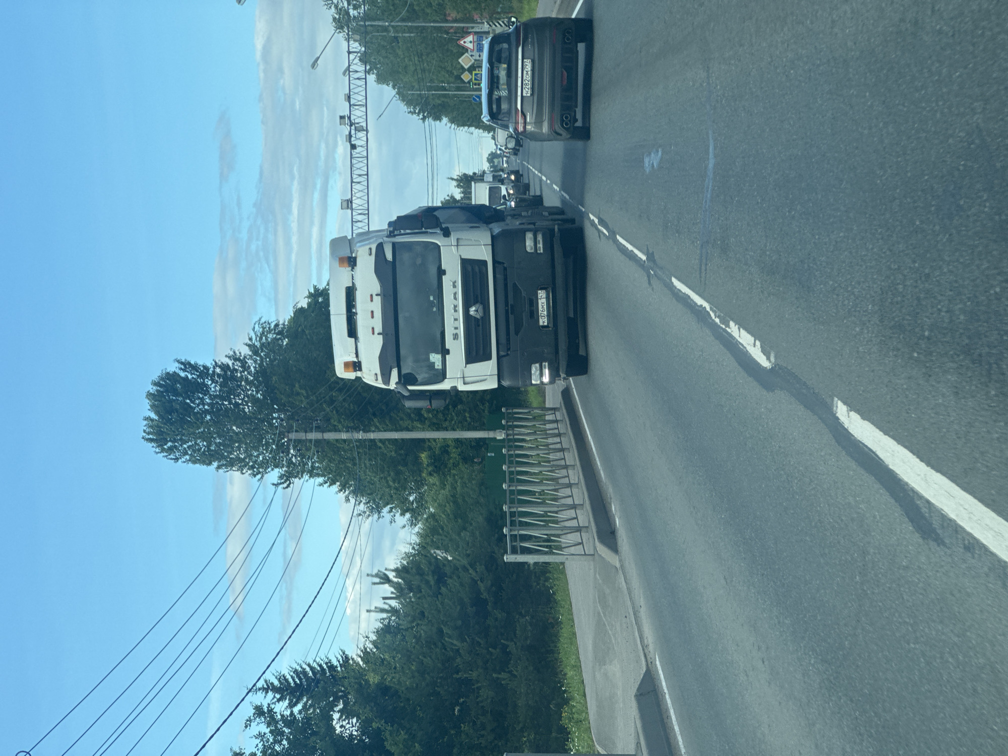 The image depicts a daytime scene on a road in Mar'ino, Russia. The primary subjects are vehicles on a highway, including a prominent white truck with "SITRAK" visible on its side. Other cars are present further down the road, suggesting traffic. The scene is set outdoors under a clear blue sky with some scattered clouds, indicating fair weather. Green trees line the side of the road, and power lines are visible overhead, typical of a semi-urban or suburban environment. There's a metal railing or fence along the edge of the road. The overall impression is of a journey or commute. Visible text includes the brand name on the truck and license plates on some of the vehicles, though the specific characters on the license plates are not clearly legible. The perspective suggests the photo was taken from inside another vehicle, possibly moving.