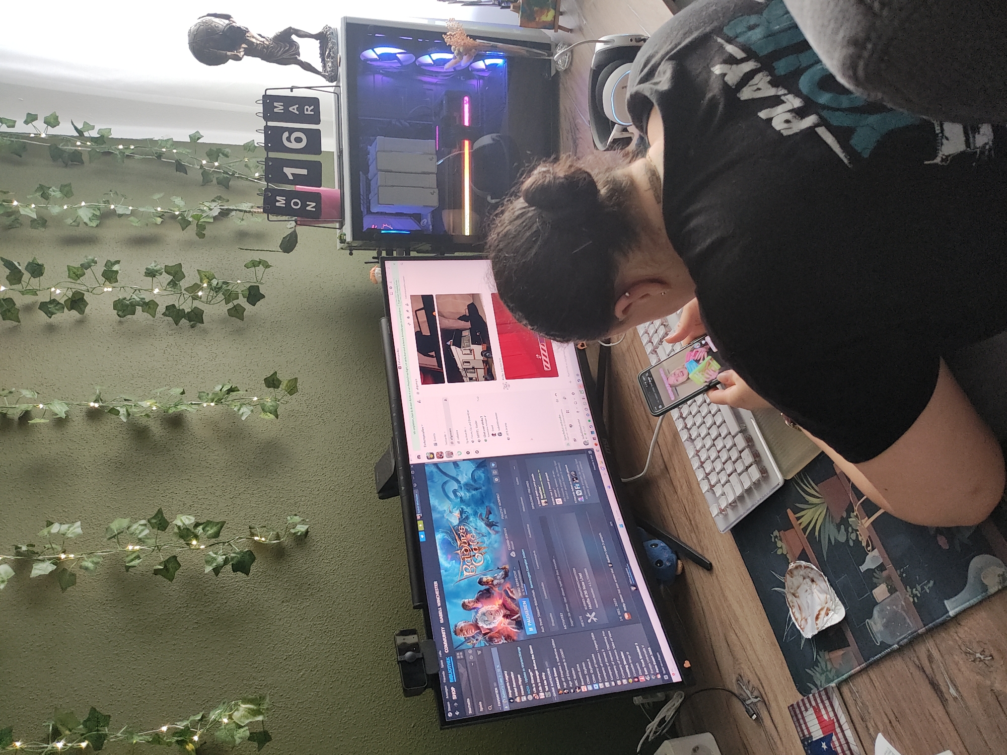 The image presents an indoor scene located in Trebgast, Germany, focused on a person seated at a computer desk setup. The individual, viewed from behind, has dark hair styled in a bun and wears a black t-shirt with partial white and teal text "PLAY YOUR" visible on the back. They are holding a smartphone in their right hand, with its screen displaying a female face with colorful hair, and appear to be actively viewing it. An earring is visible in the person's left ear.
The desk is constructed of wood. On its surface, a large ultra-wide monitor centrally displays a gaming platform interface, specifically Steam, with "Baldur's Gate 3" prominently featured and other game titles listed in German. A webcam is mounted on top of this monitor. To the right of the ultra-wide monitor, a second, smaller monitor is partially visible. A white mechanical keyboard with light-colored keycaps is positioned in front of the monitors. To the left of the keyboard, a mousepad featuring a plant motif holds a white, empty seashell. Adjacent to the mousepad is a small rectangular item displaying a Texas flag design.
To the right of the monitors, a transparent computer tower is visible, showcasing internal components illuminated by blue and pink RGB lighting from fans and other hardware. On top of the computer tower rests a flip calendar displaying "MON 16 MAR" and a small pink container holding a green plant. Further to the right, a figurine depicting Atlas holding a globe on his shoulders is present.
The wall behind the desk is textured and painted an olive-green color. It is adorned with multiple vertical strands of artificial ivy, each interspersed with small, illuminated fairy lights. A bright white curtain or window is partially visible on the far right side of the image, suggesting a source of natural light. The person is seated in a grey upholstered chair, parts of which are visible.